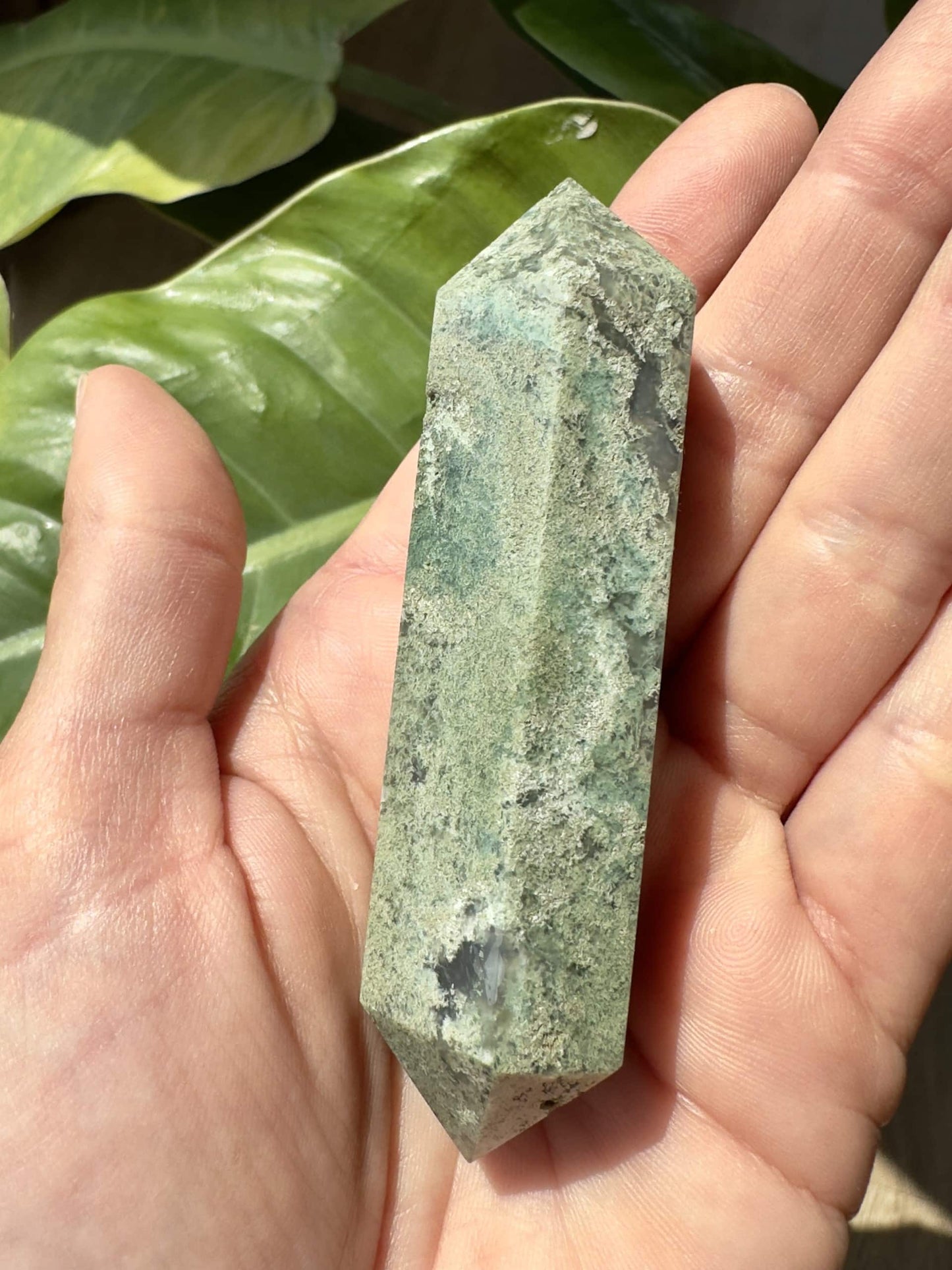 En dobbelt termineret spids i Mos Agat holdes i en hånd under sollys med grønne blade i baggrunden.