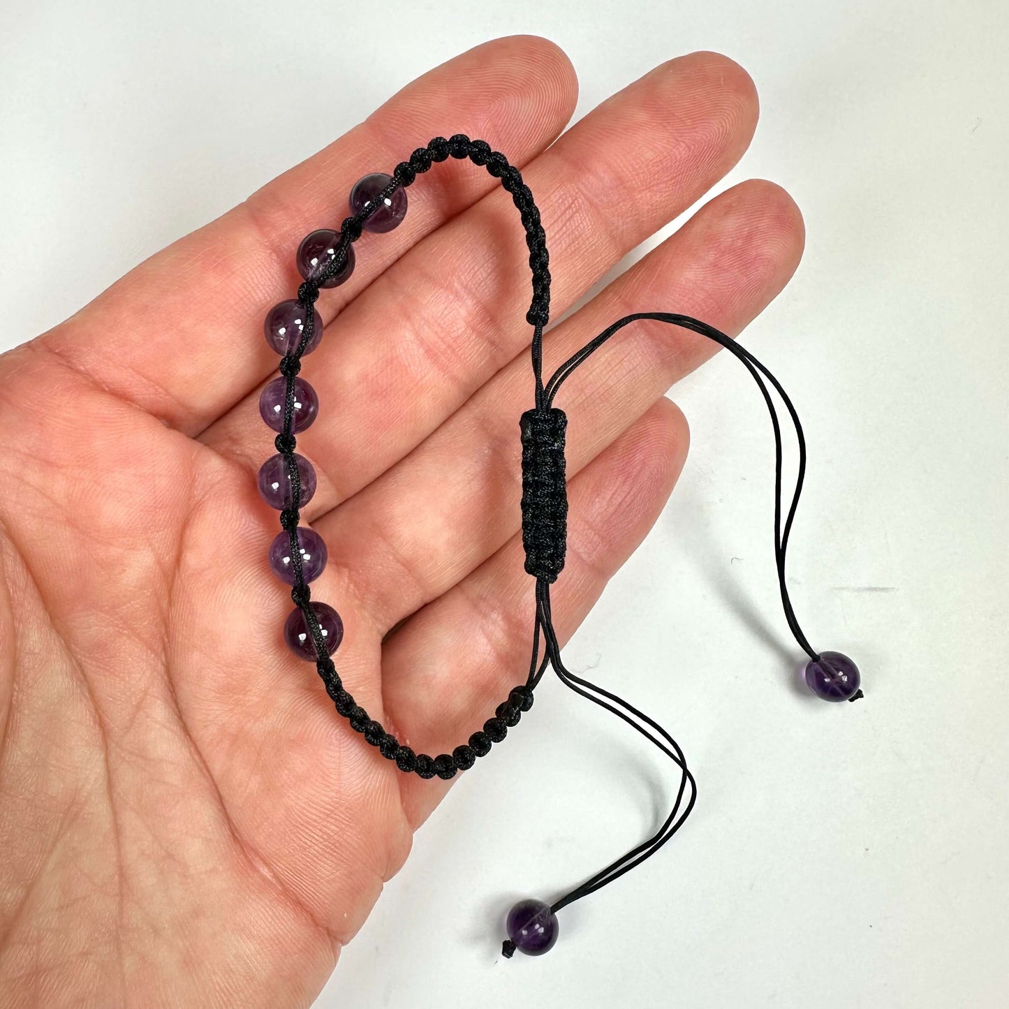 Hånd holder et ametyst armbånd med sorte snore og lilla perler.