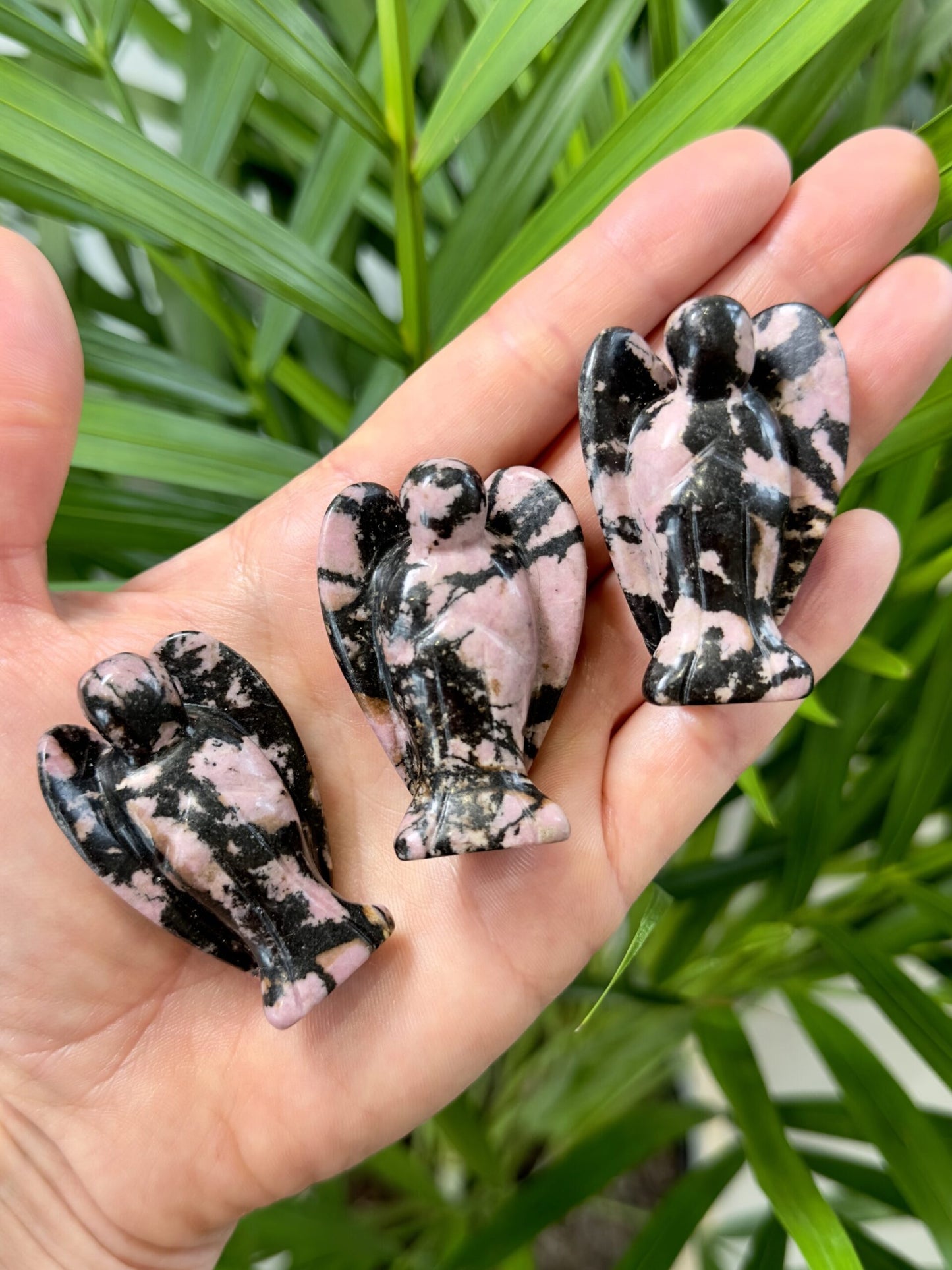 Hånd holder tre små rhodonit engle foran grønne blade i baggrunden.