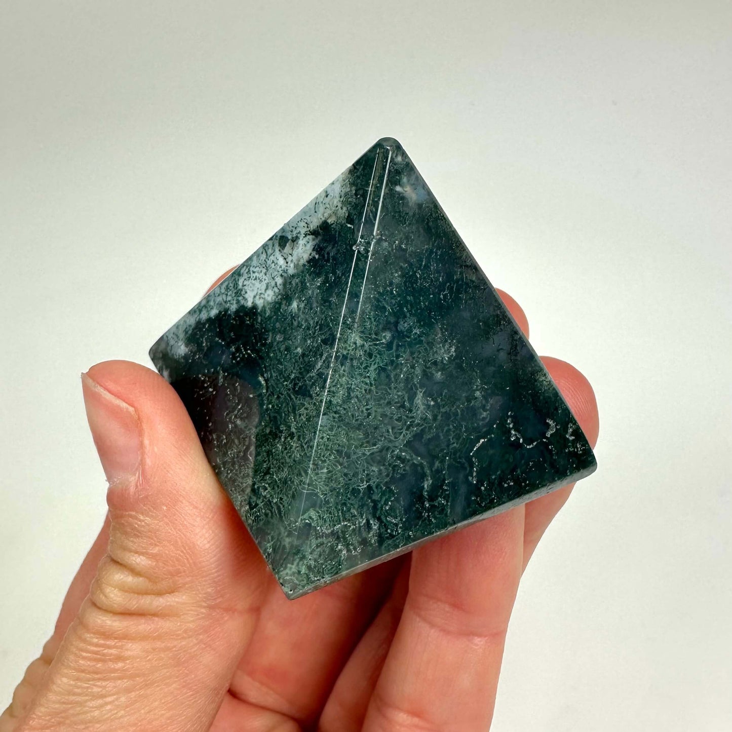 Hånd holder en mos agat pyramide med grønne og mørke nuancer på en lys baggrund.