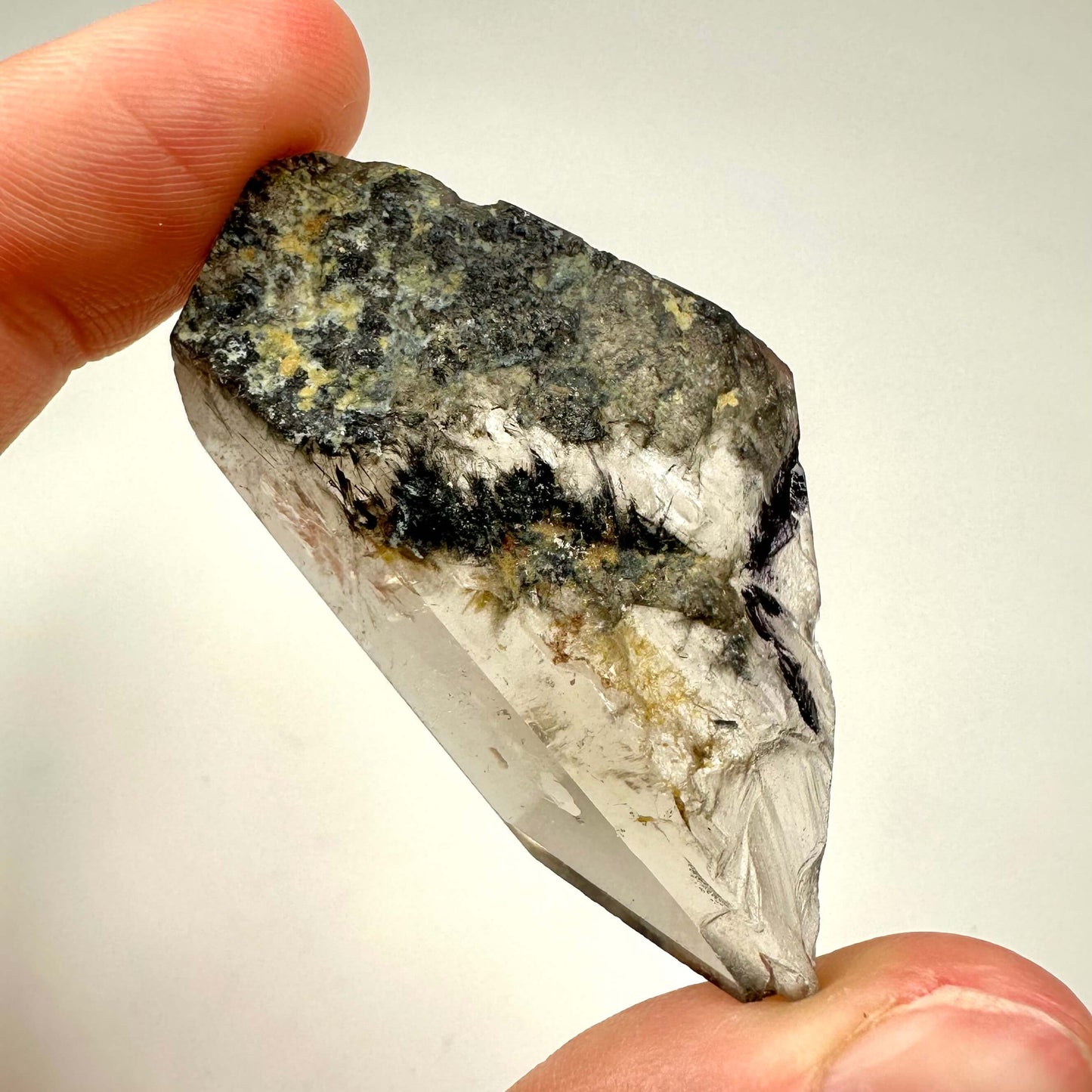 Fantomkvarts fri form holdes mellem fingre med en lys baggrund. Mørke og lyse nuancer på krystaloverfladen.