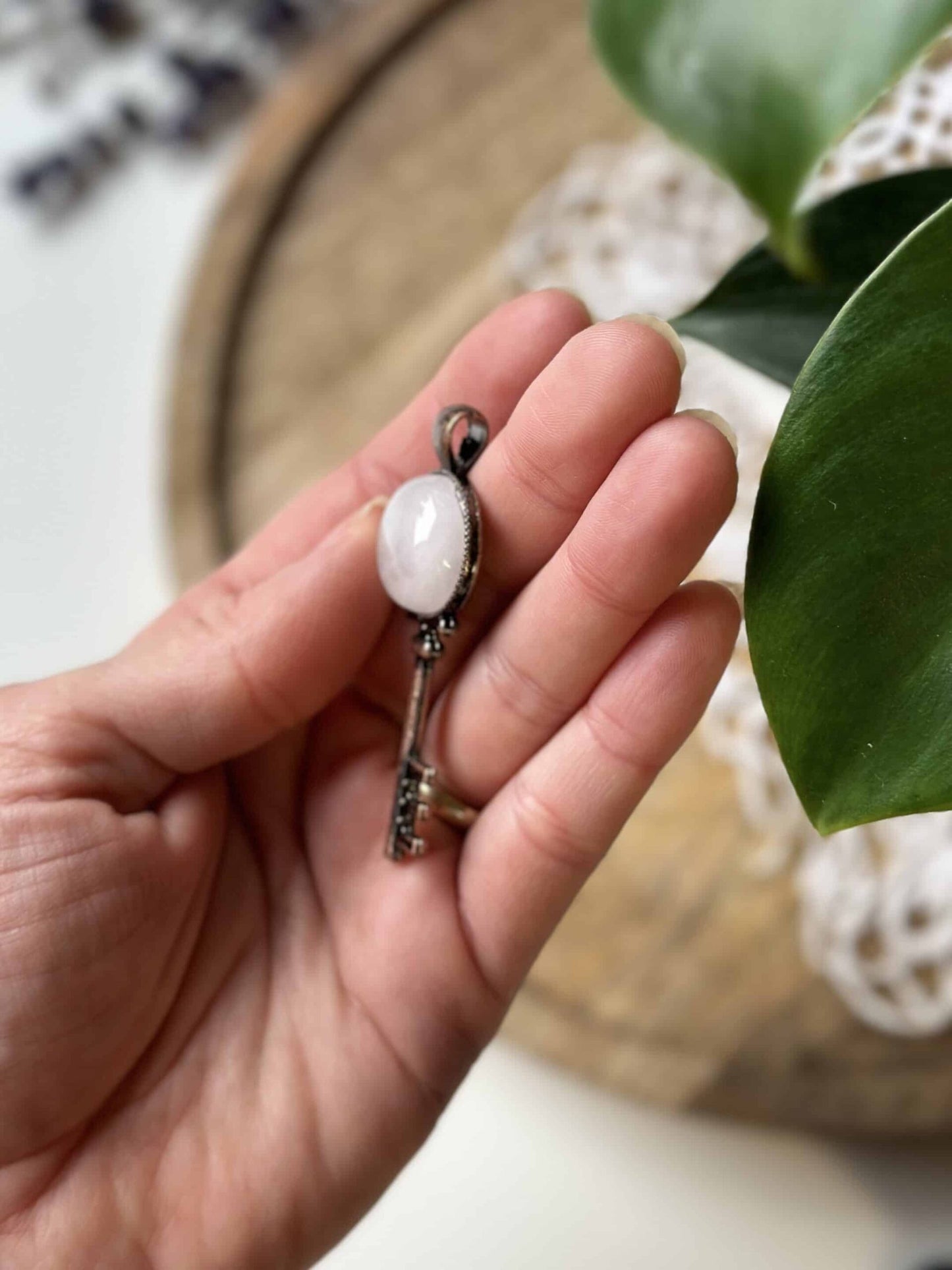 Hånd holder en dekorativ nøgle med rosenkvarts vedhæng, med grøn plante i baggrunden.