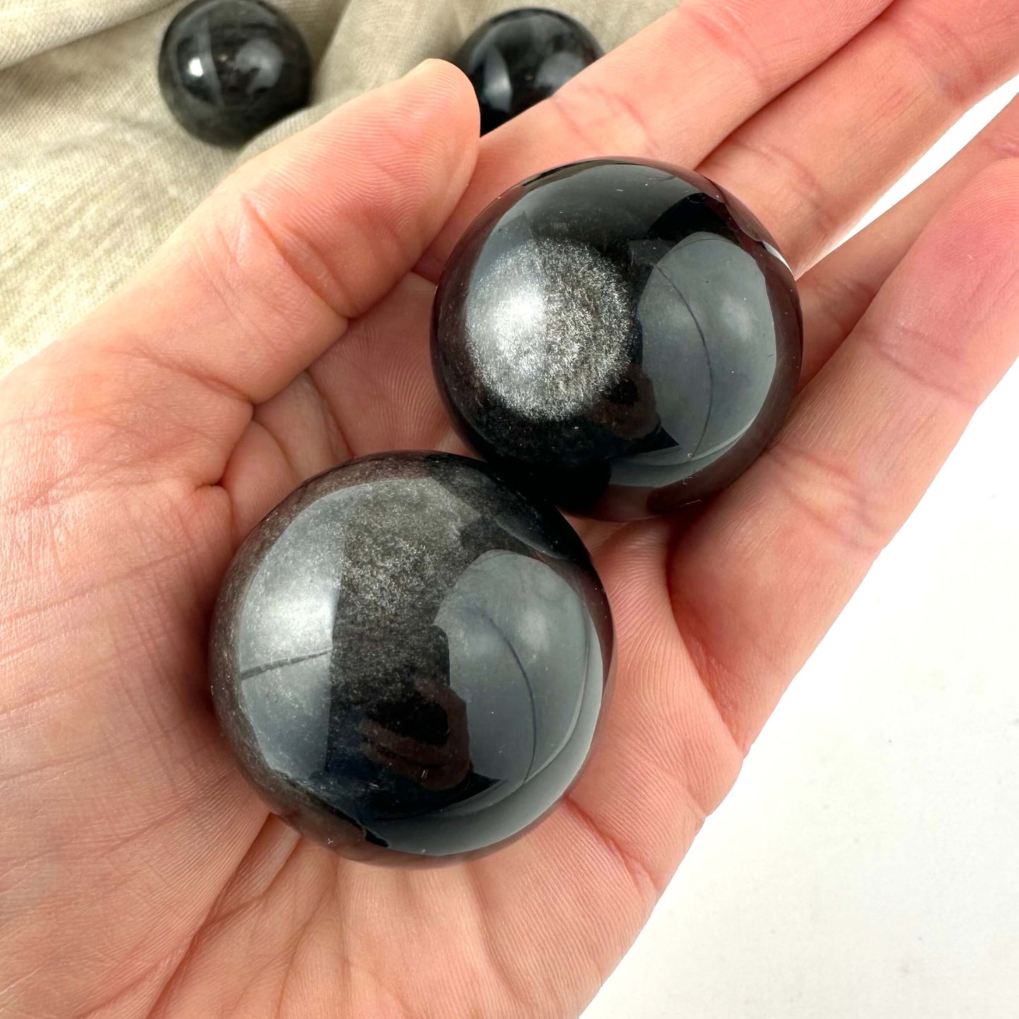 Hånden holder to glatte Obsidian gylden kugler med reflekterende overflade. En beige baggrund ses delvist.