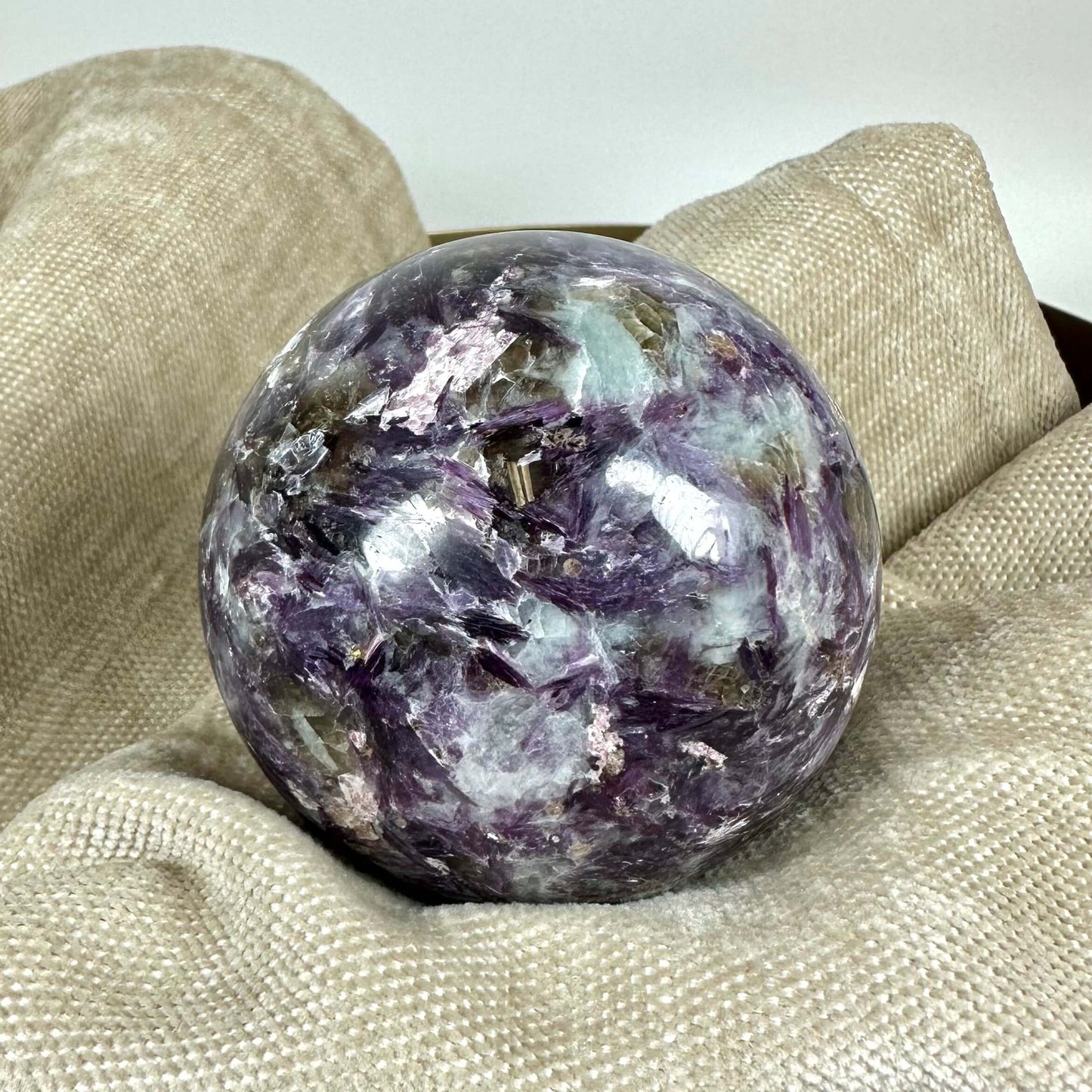 Lepidolit kugle med lilla og beige nuancer ligger på beige tekstilbaggrund.
