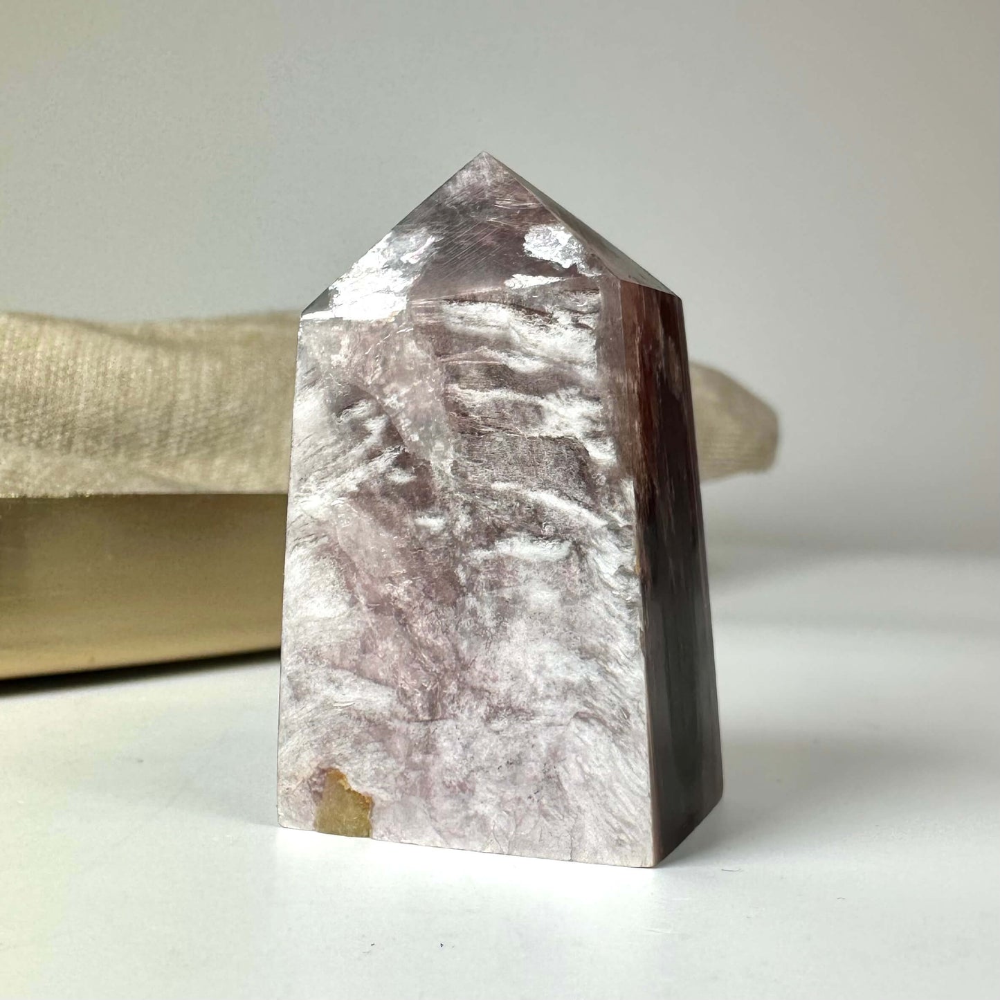 En poleret Lepidolit Mica-krystal med en pyramideformet top og varieret lilla farvenuance står på en lys overflade.