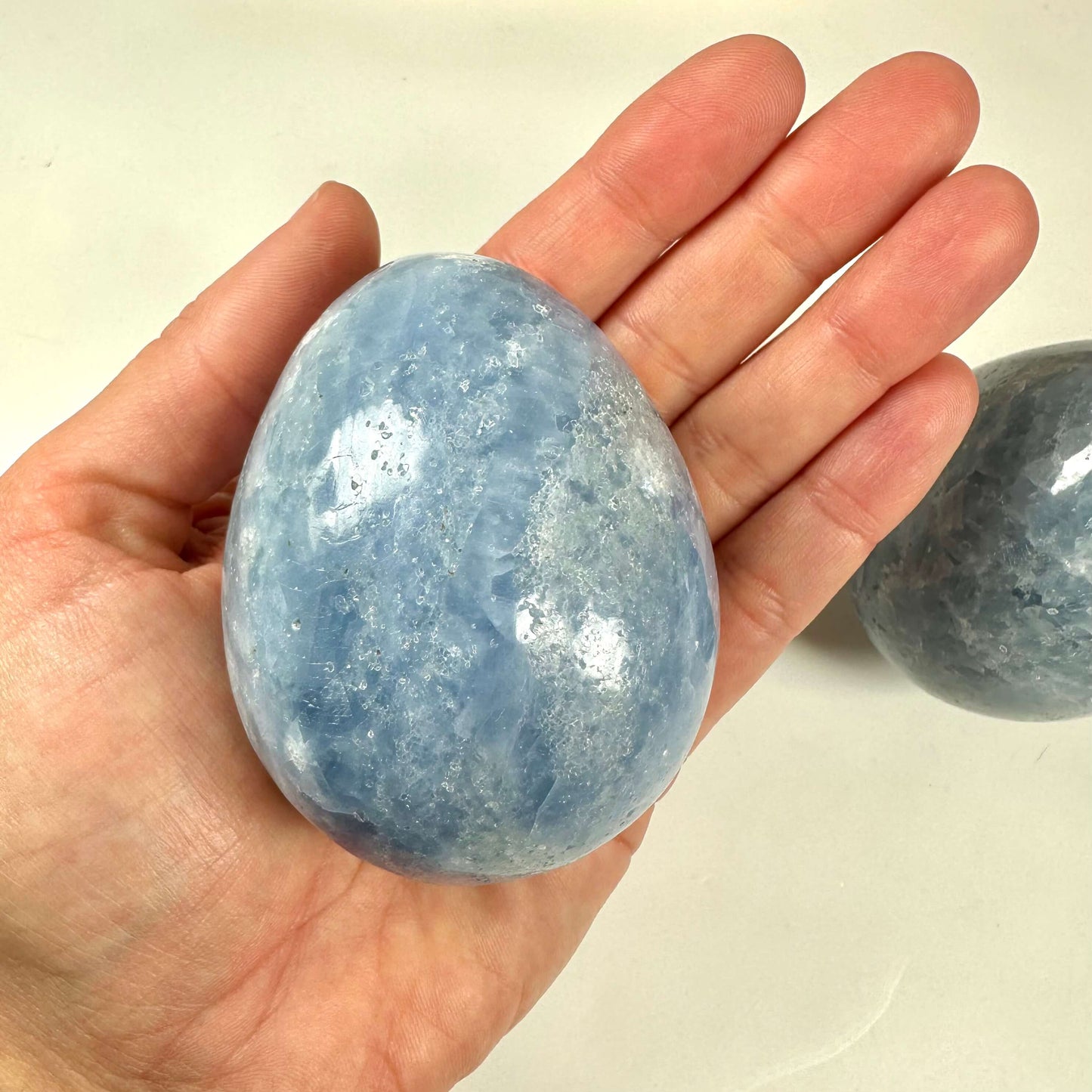 En hånd holder et stort, glat blå calcit æg, mens et andet æg ses i baggrunden.