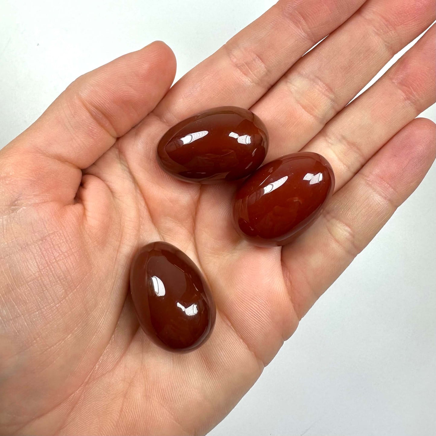 En hånd, der holder tre glatte, brunrøde karneol æg mod en lys baggrund.