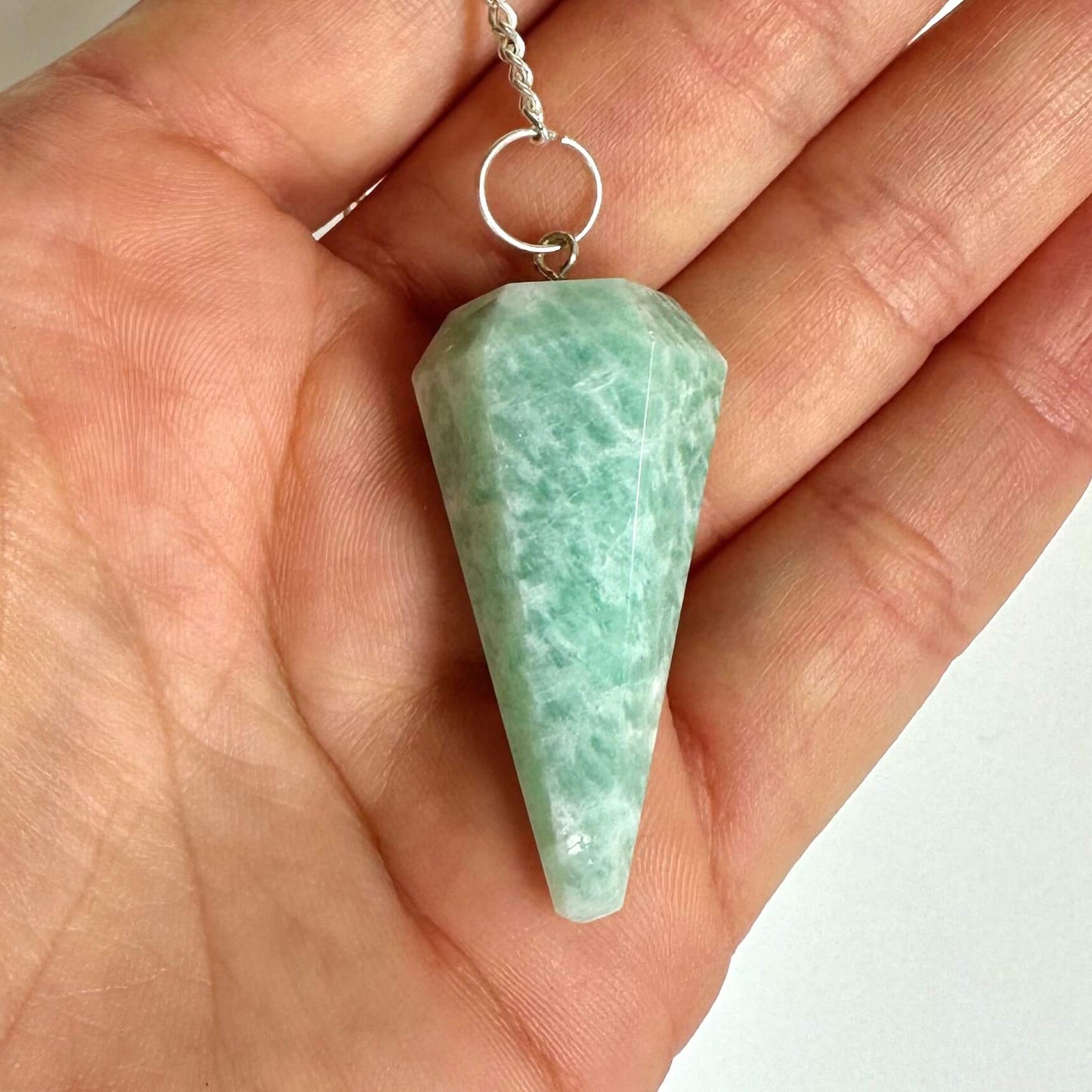 Person holder et grønt amazonit pendul i hånden, med en sølvkæde fastgjort til pendulet.