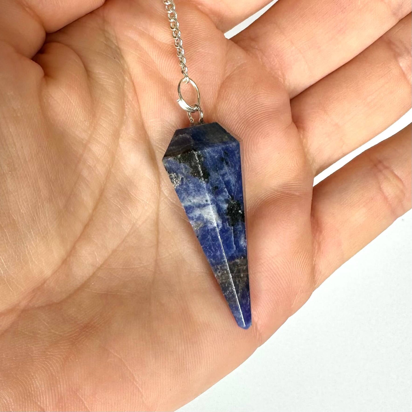 En hånd holder et blåligt Sodalit pendul med en sølvkæde.