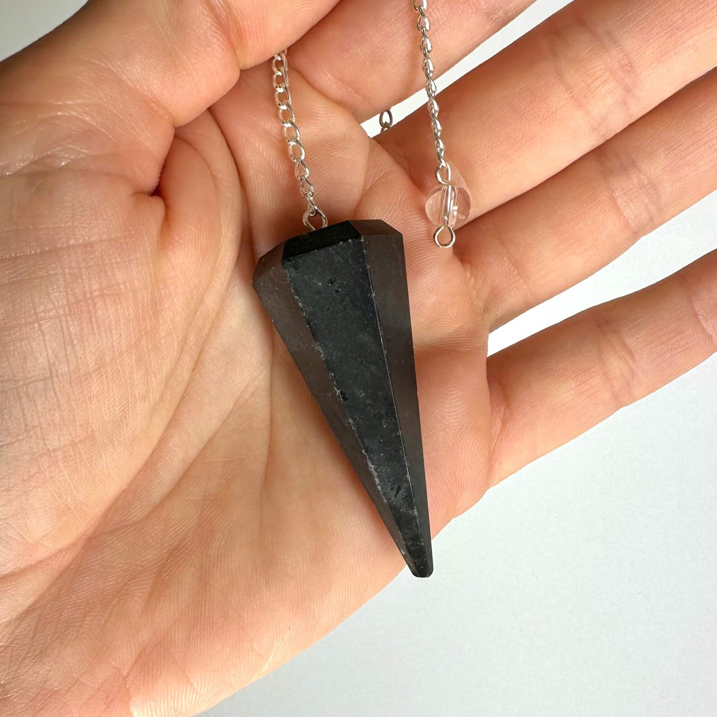 En hånd holder et shungit pendul med en sølvkæde, tæt op mod en lys baggrund.