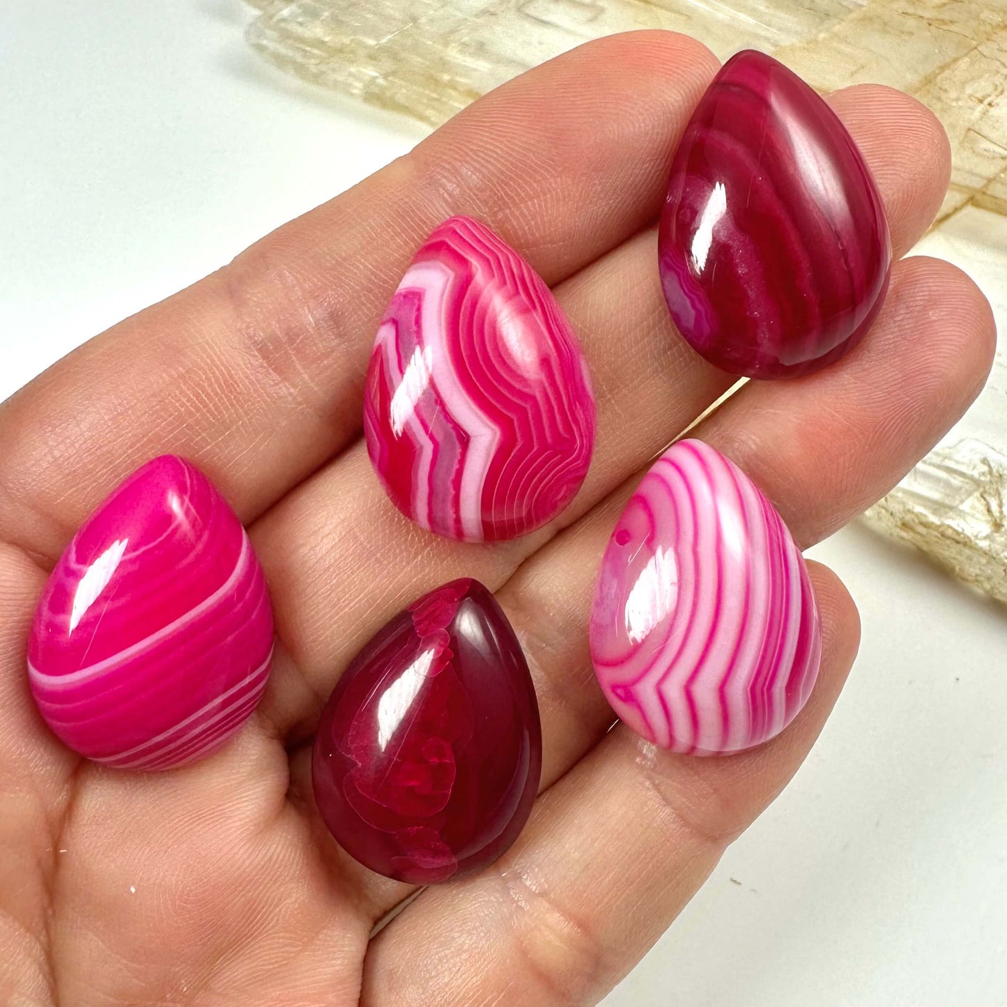 Hånd med fem pink agat dråbeformede sten med mønster og nuancer i pink og hvid.