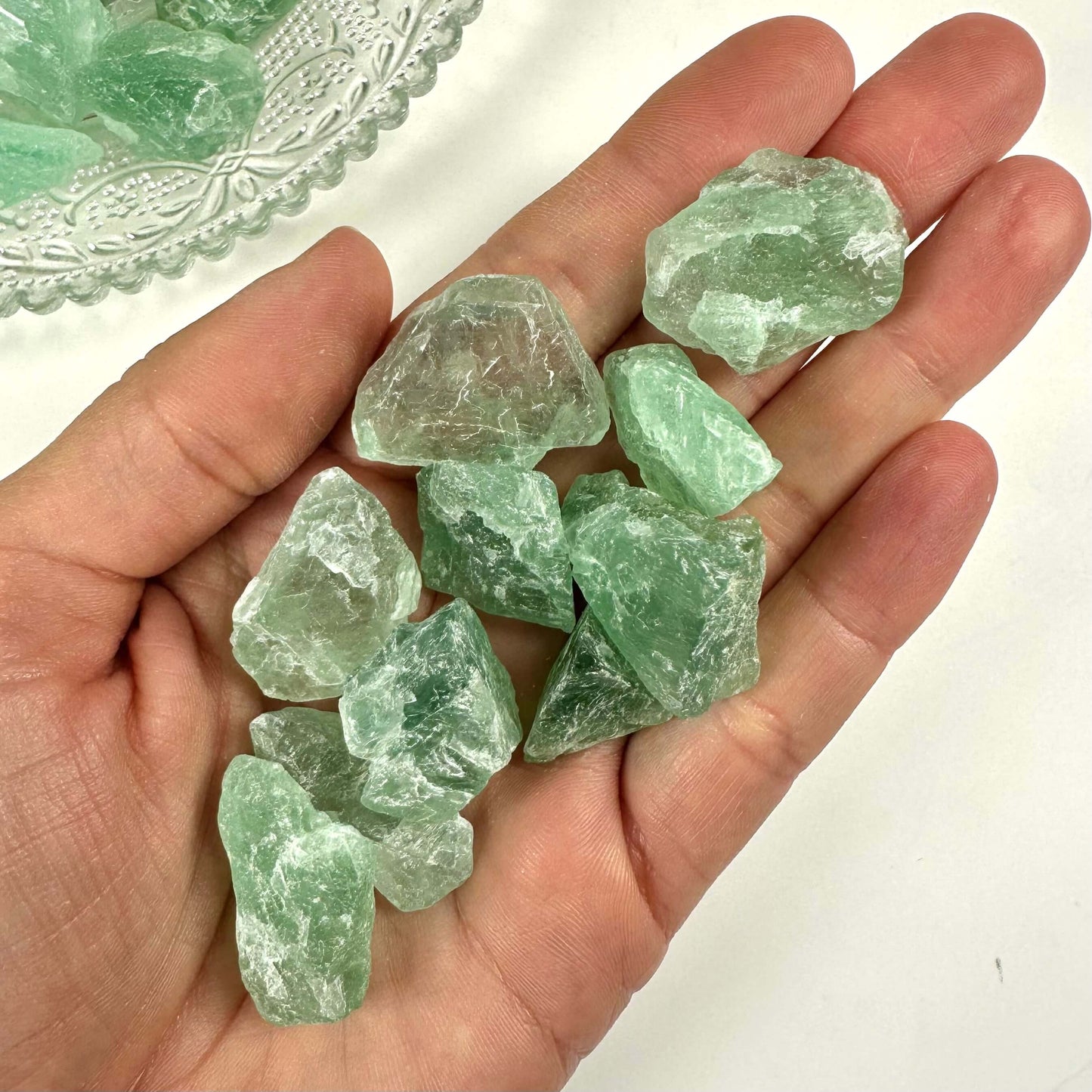 Hånd holder små stykker grøn fluorit rå, placeret over en hvid baggrund. En tallerken med flere sten ses i baggrunden.