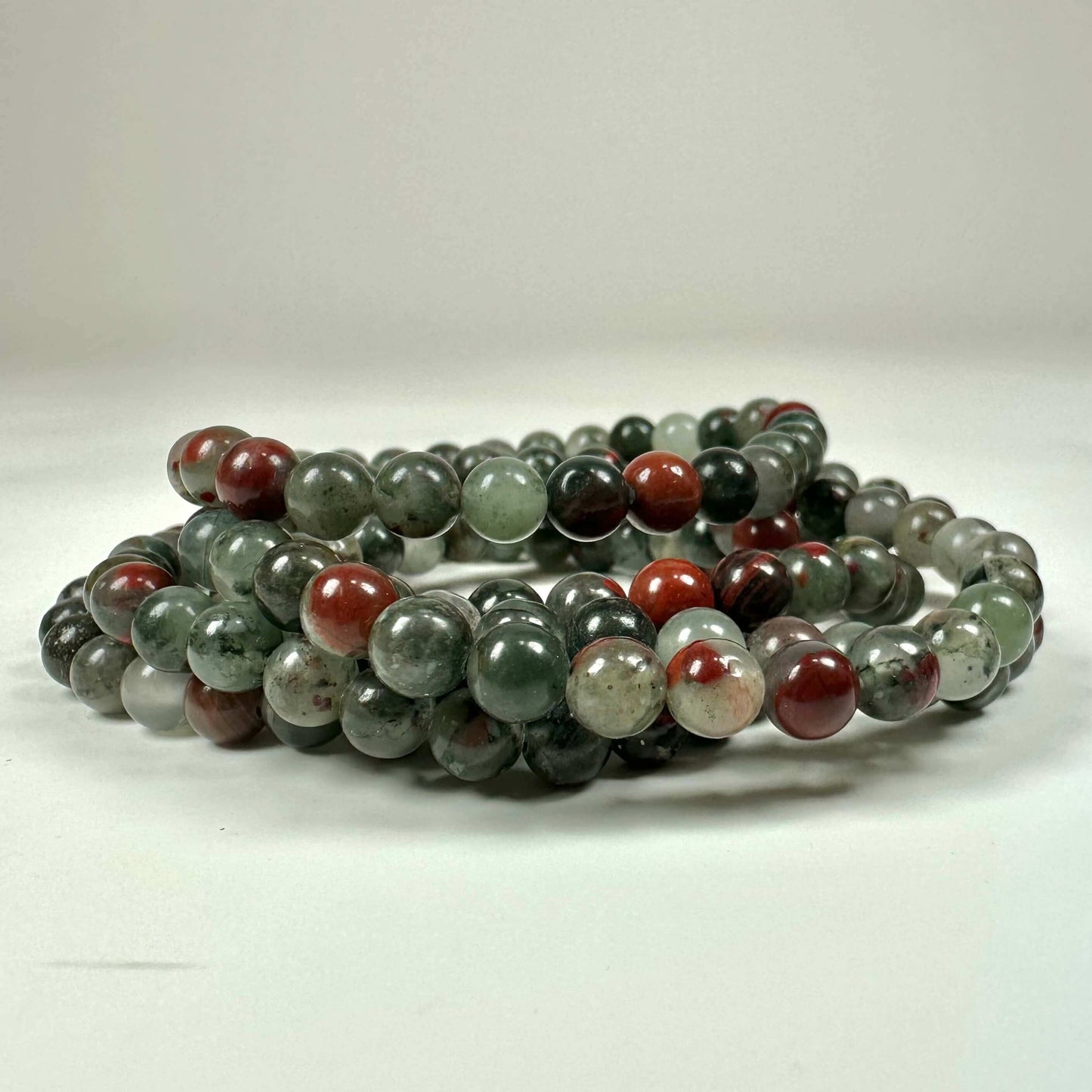 Armbånd af flerfarvede stenperler i nuancer af grønt, rødt og gråt i en snoning på hvid baggrund.