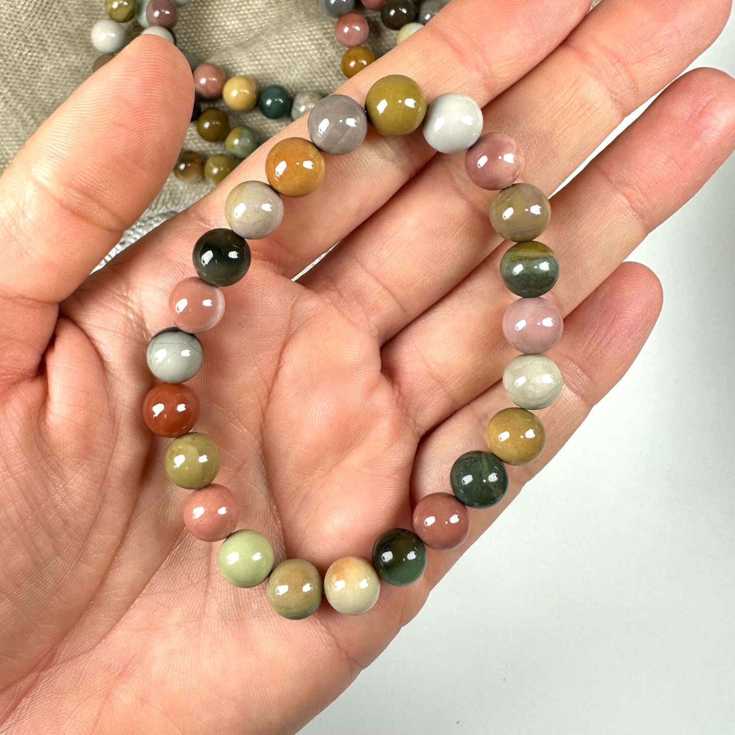 En hånd holder et armbånd med farverige, blanke stenperler på en lys baggrund.