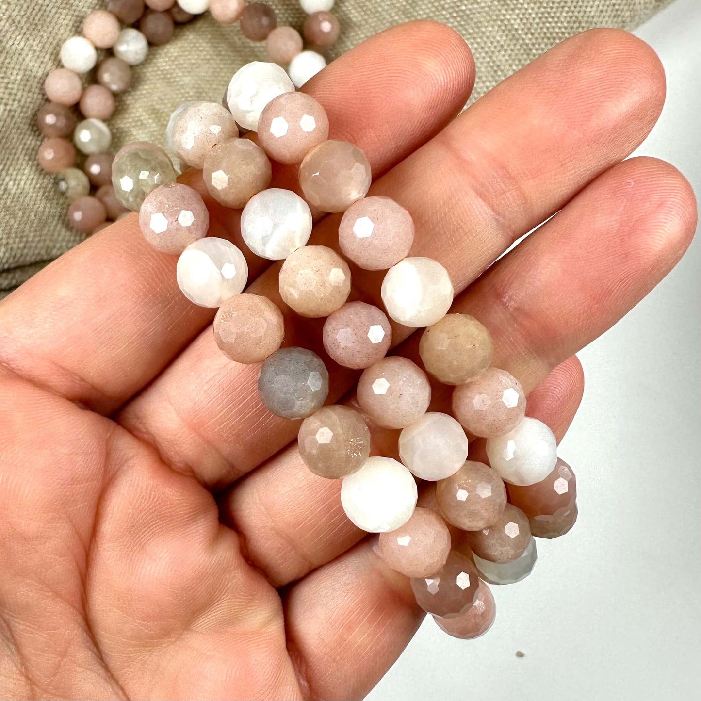 Hånd holder en række Månesten mix armbånd med facetterede perler i nuancer af hvid, beige og rosa.