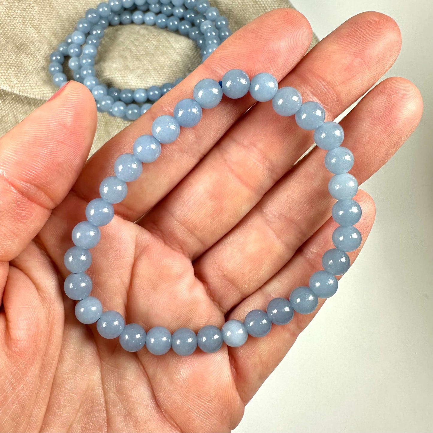 Hånd der holder et Angelit armbånd af lyseblå perler, med lignende armbånd i baggrunden på en lys overflade.
