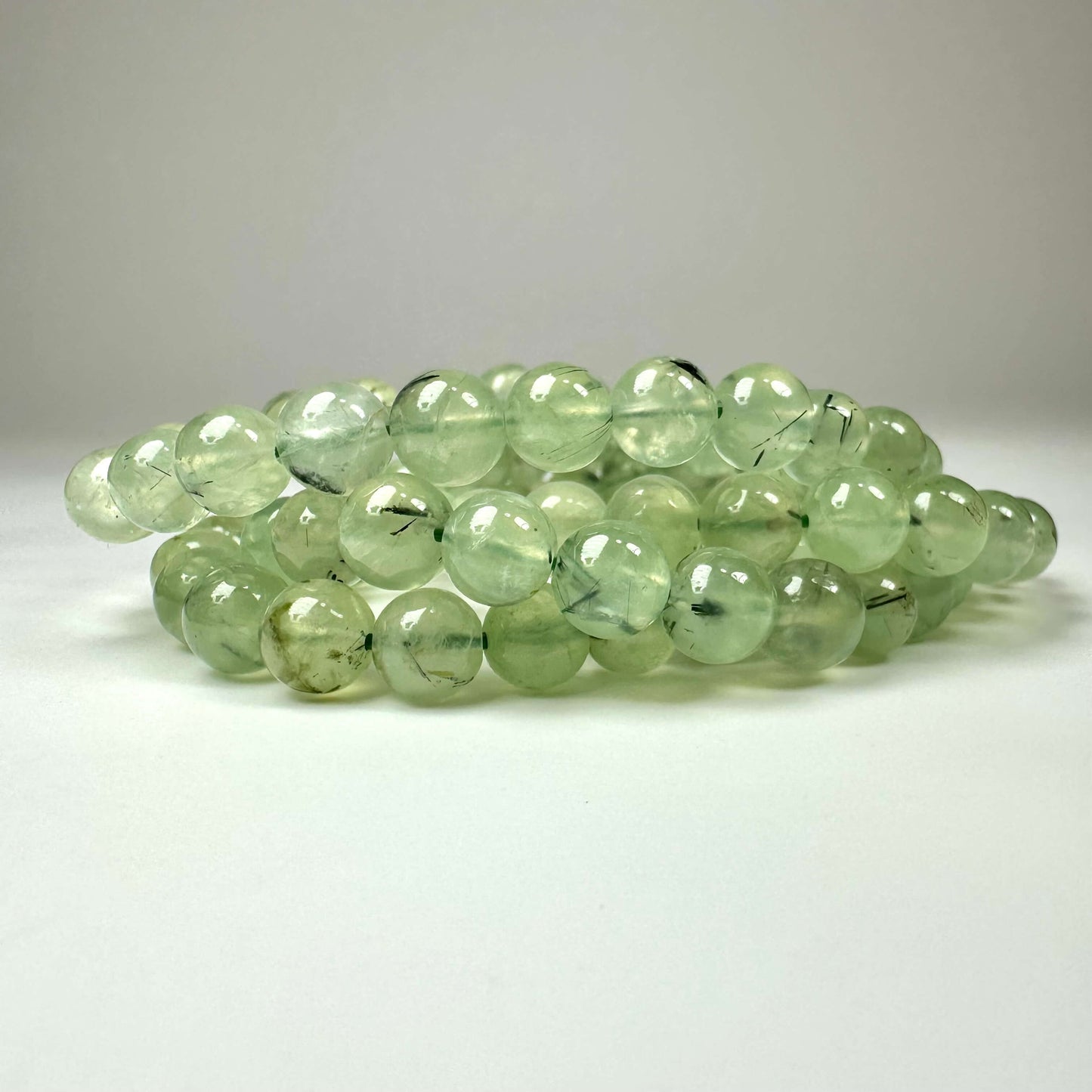 Prehnit armbånd 8mm med lysegrønne, glansfulde perler lagt ud på en lys baggrund.