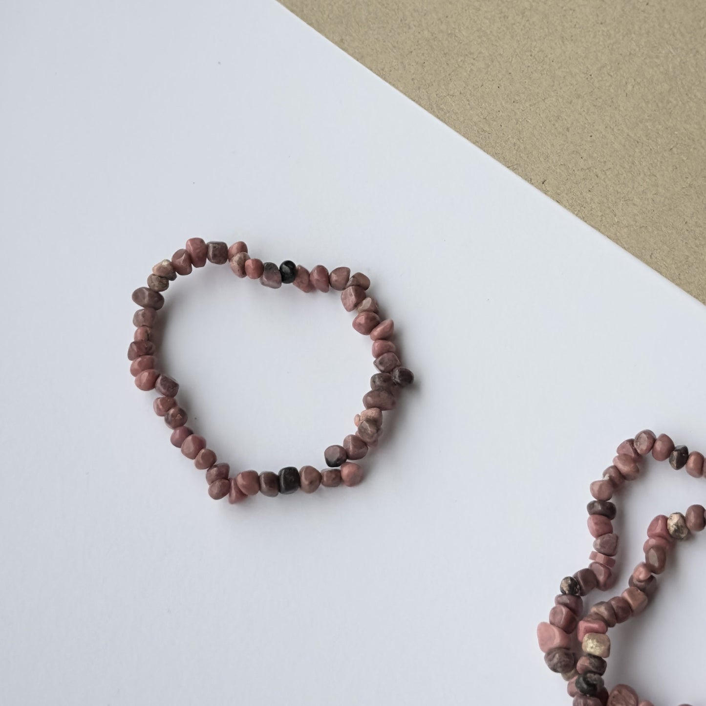 Et Rhodonit armbånd ligger på en hvid baggrund med en beige overflade i øverste hjørne. Armbåndet er lavet af små, ujævne rosa og sorte sten, der symboliserer kærlighed, følelsesmæssig balance og heling. Stenene har naturlige nuancer og struktur, der fremhæver deres unikke udtryk. Et andet Rhodonit armbånd ses delvist i billedets højre hjørne.