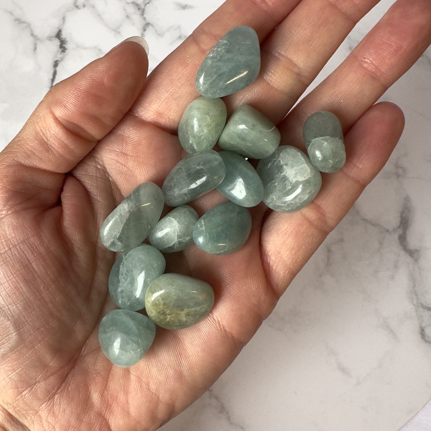 En hånd holder flere glatte, polerede akvamarin lommesten i nuancer af lyseblå og havgrøn. Stenene varierer lidt i størrelse og form og har en glasagtig, skinnende overflade. Baggrunden er en hvid marmorflade, som fremhæver stenens blågrønne farve. Akvamarin siges at have beroligende og styrkende egenskaber og forbindes ofte med indre fred og kommunikation.