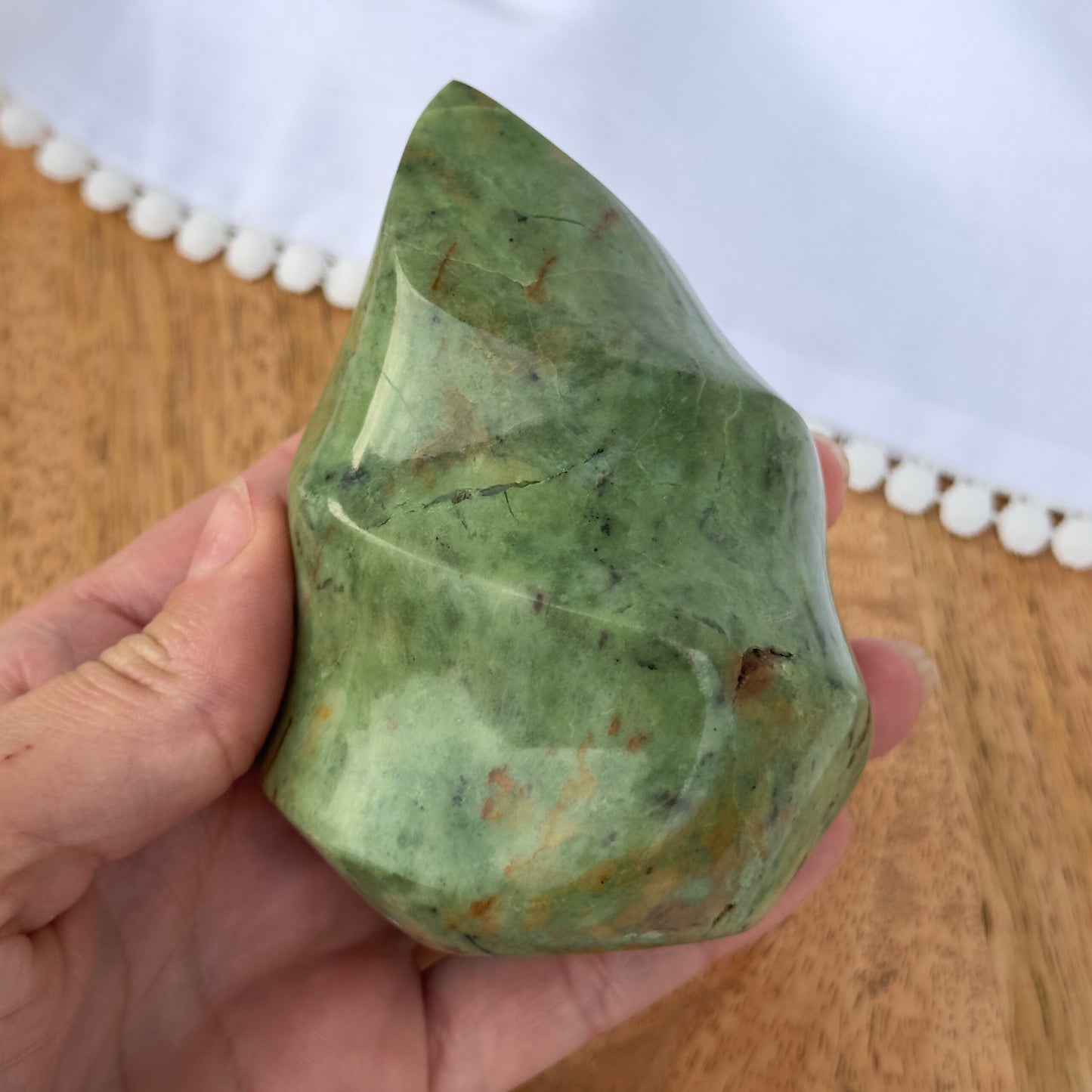 Hånd holder en poleret, grøn Chrysopras flamme på træbord med hvid baggrund.