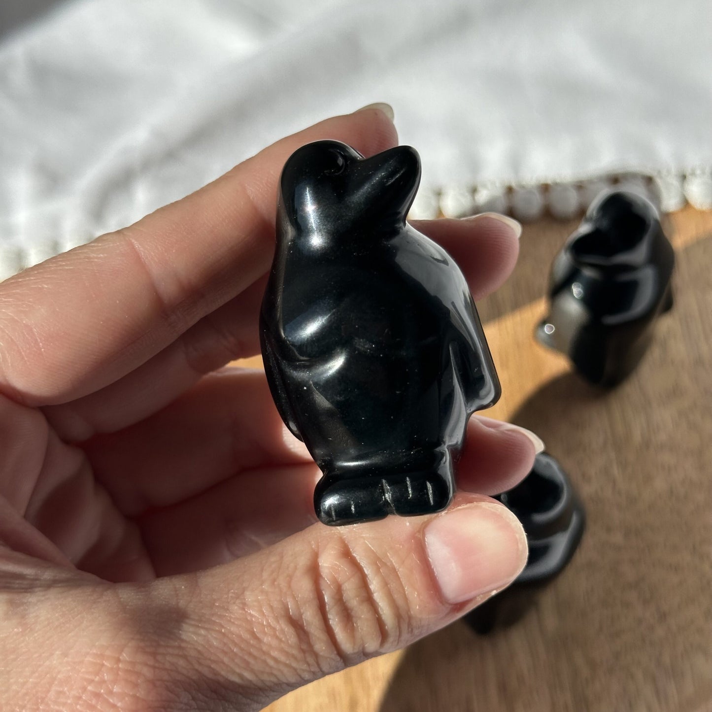 Hånd holder en lille skulptur af en obsidian pingvin, med bord og andre figurer i baggrunden.