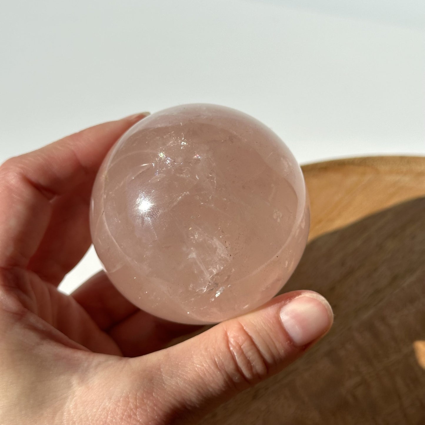 Hånd holder en rosenkvarts kugle, der lyser i sollys. En træoverflade anes i baggrunden.