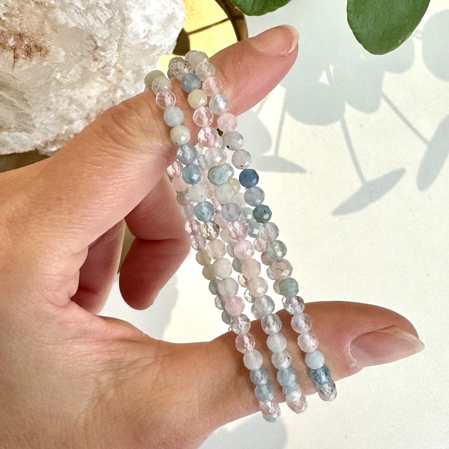 En hånd holder flere farverige Morganit armbånd i lyset mod en lys baggrund.