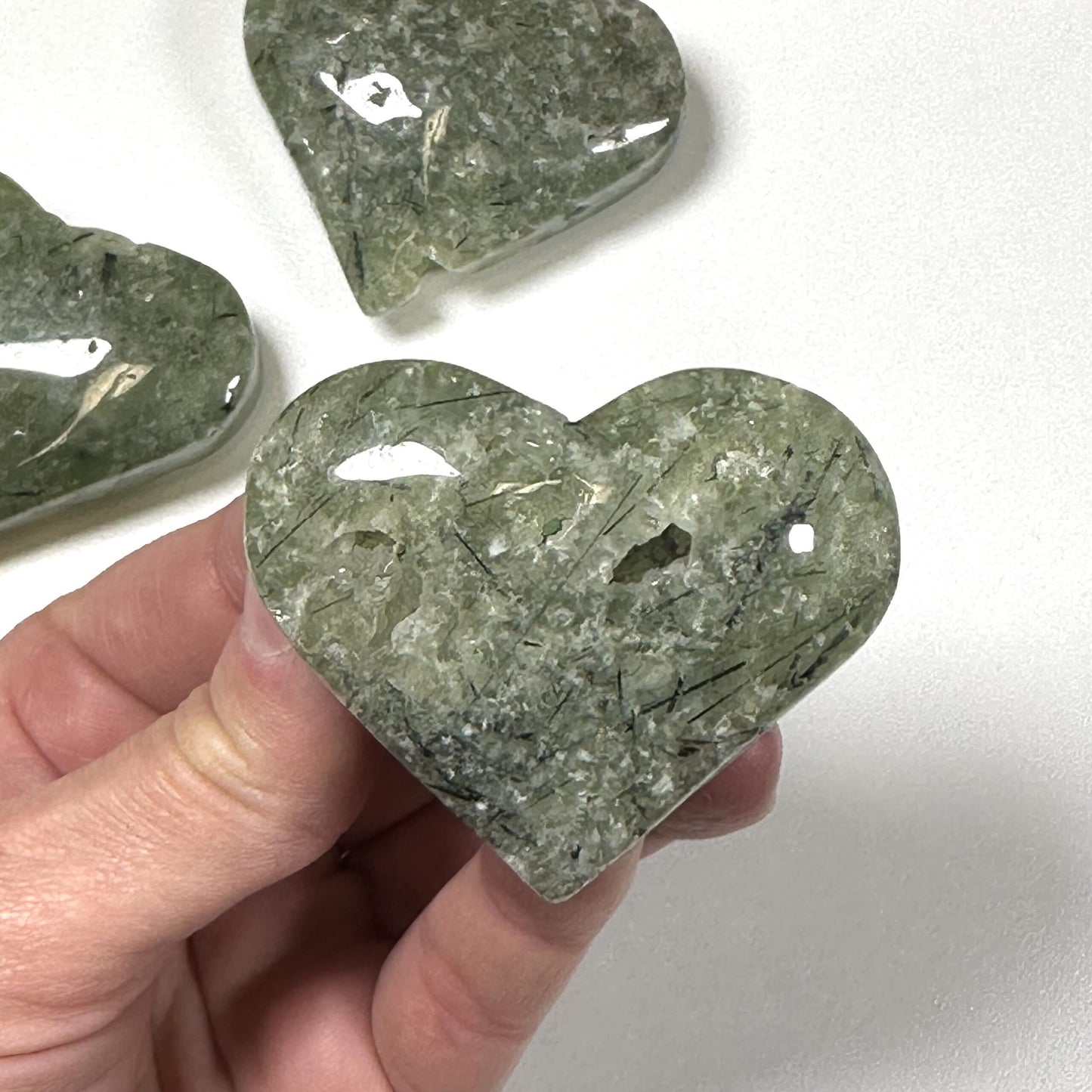 Hånd holder et poleret Prehnit hjerte med to andre i baggrunden på en hvid overflade.