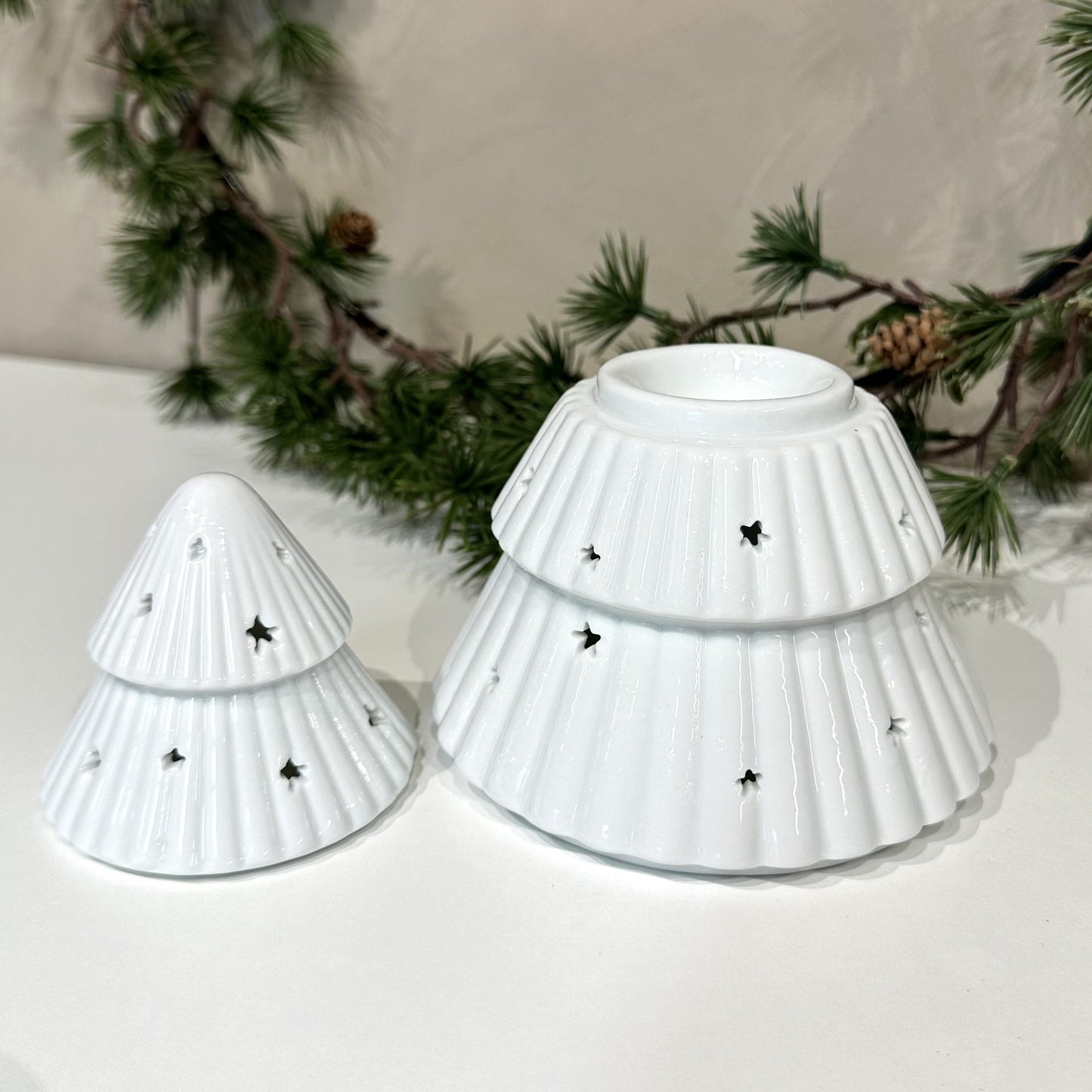 To hvide Voksbrænder juletræer med stjerneformede åbninger på en hvid overflade, omgivet af grankviste.