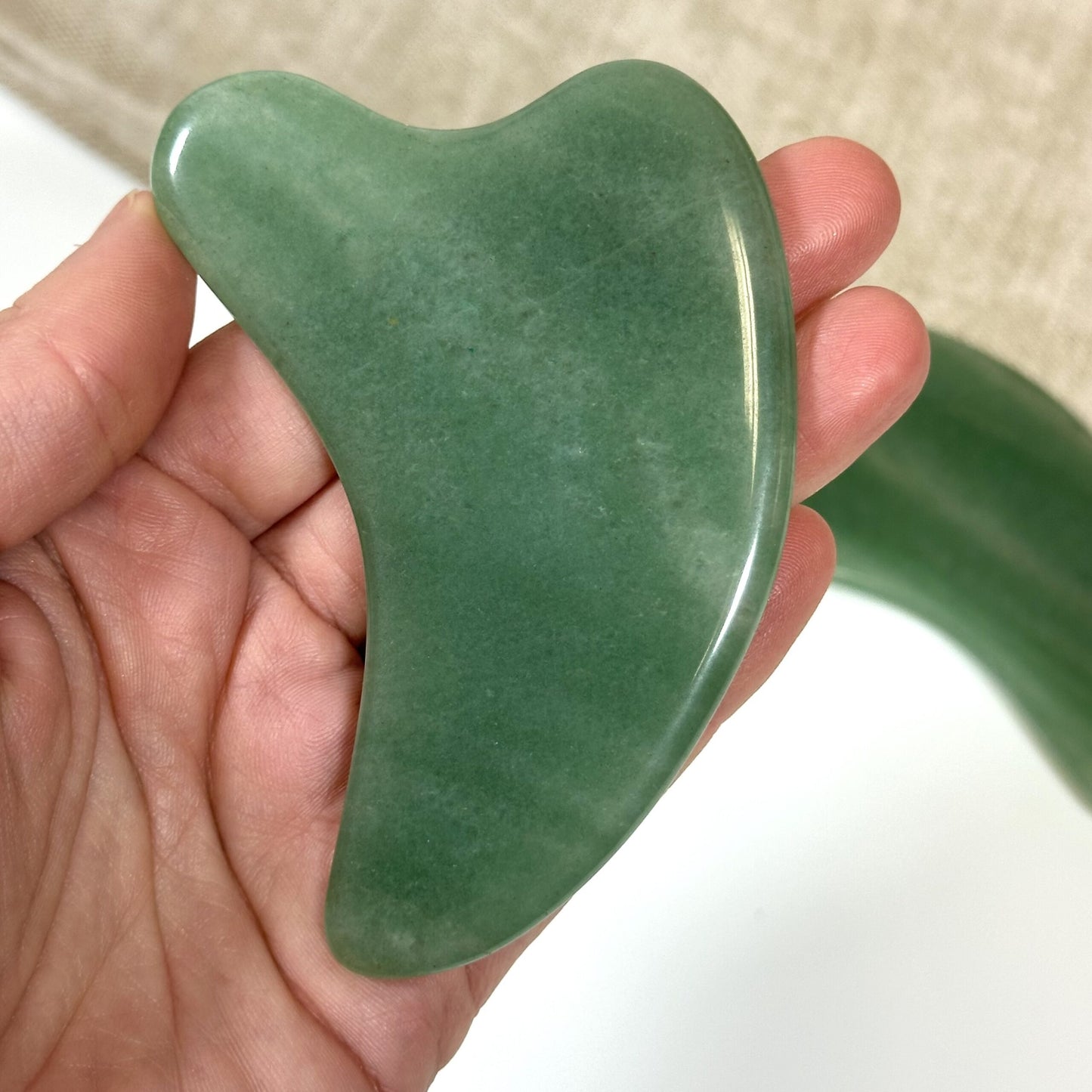Hånd holder en grøn Aventurin Gua Sha, formet til ansigtsmassage, på en lys baggrund.
