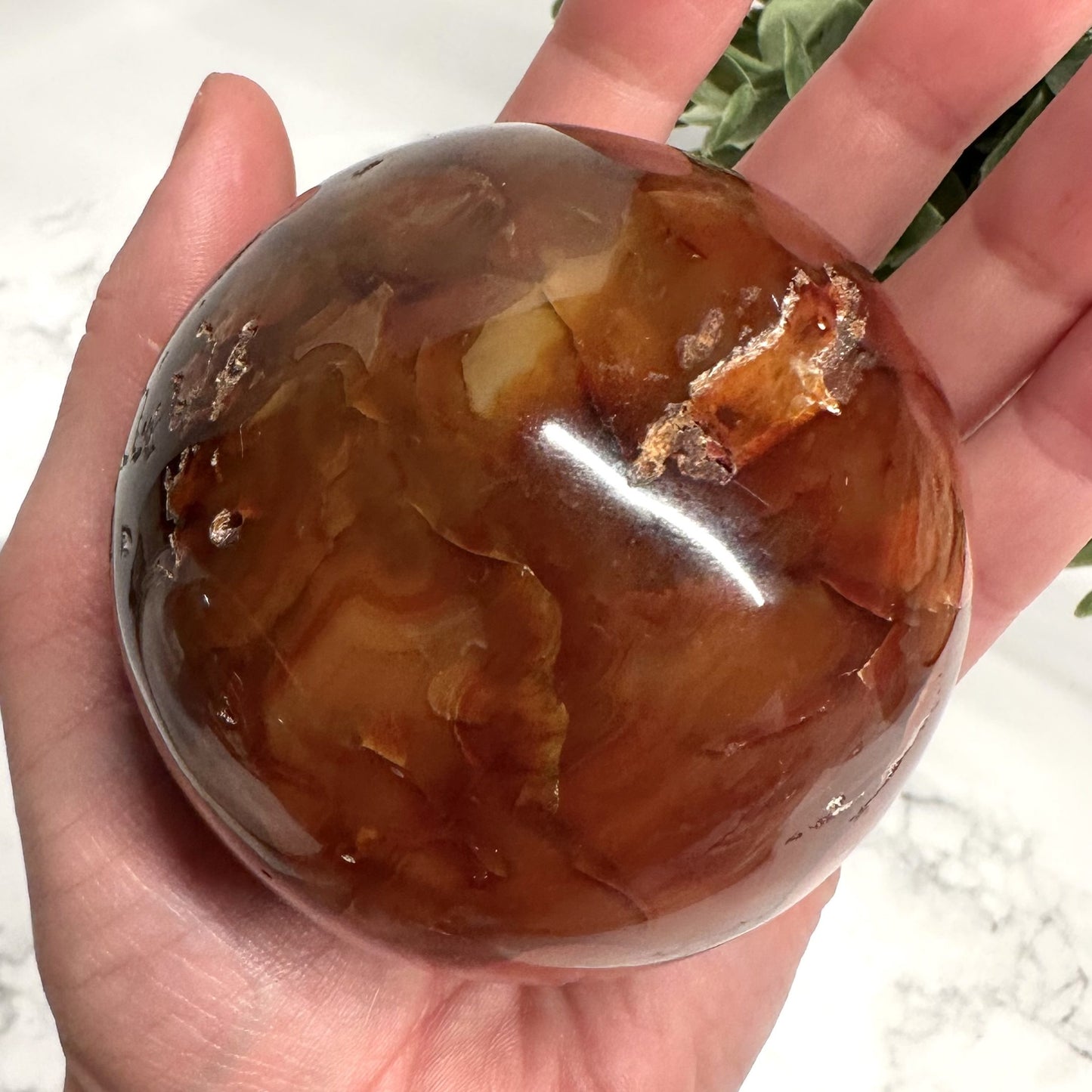 En hånd holder en glat, poleret karneol kugle med dybe brune og gyldne nuancer.