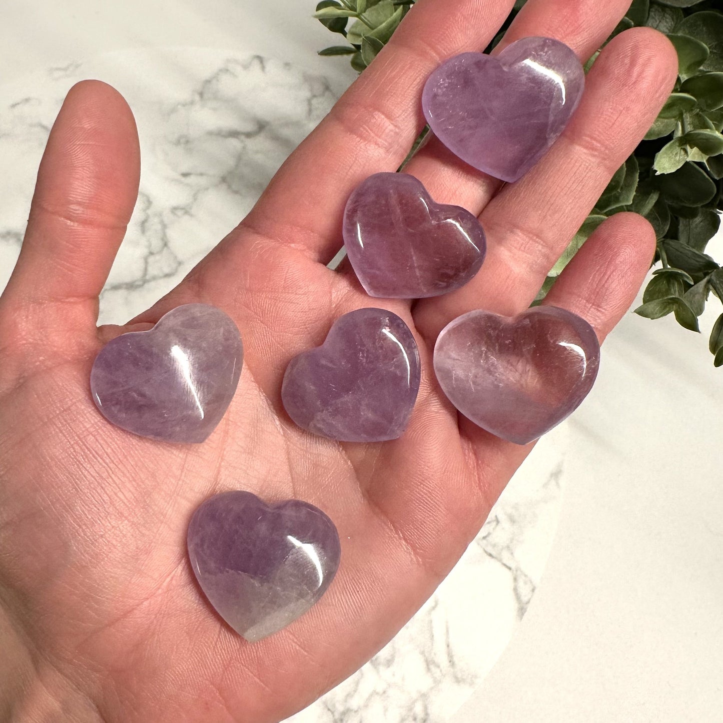 Hånd holder seks ametyst hjerter på en marmorbaggrund med grønne blade i baggrunden.