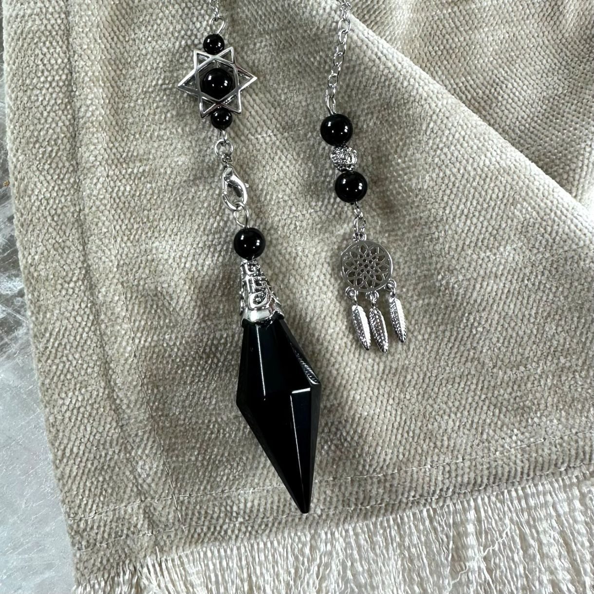 Sort obsidian pendul med stjerne og drømmefanger detaljer, placeret på en beige stofbaggrund.