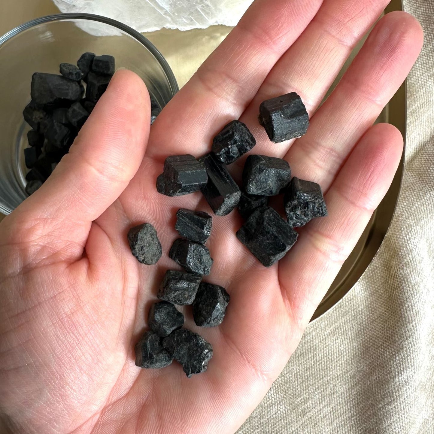 Håndfuld sort turmalin mini stave vises. Flere sorte krystaller i en glasskål ses i baggrunden.