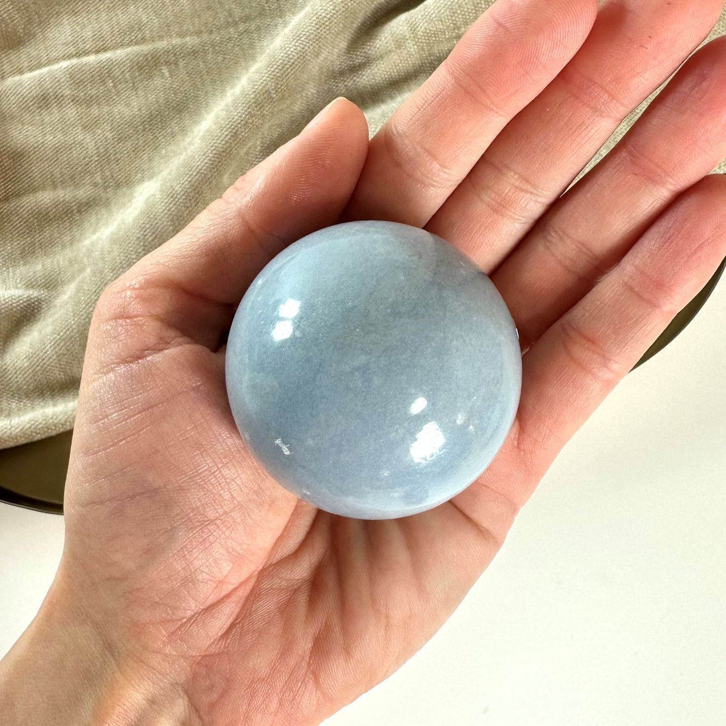 Hånd holder en glat, lyseblå Angelit kugle oven på en beige klud.