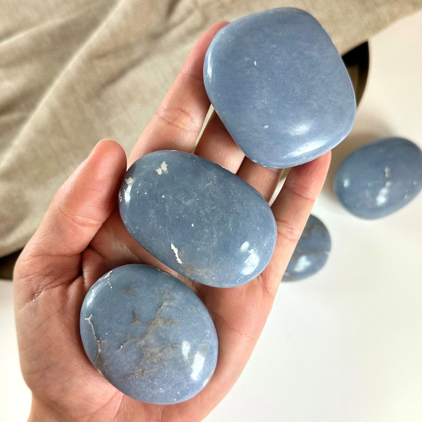 Hånd holder tre glatte, blå Angelit palmstones mod en beige baggrund.