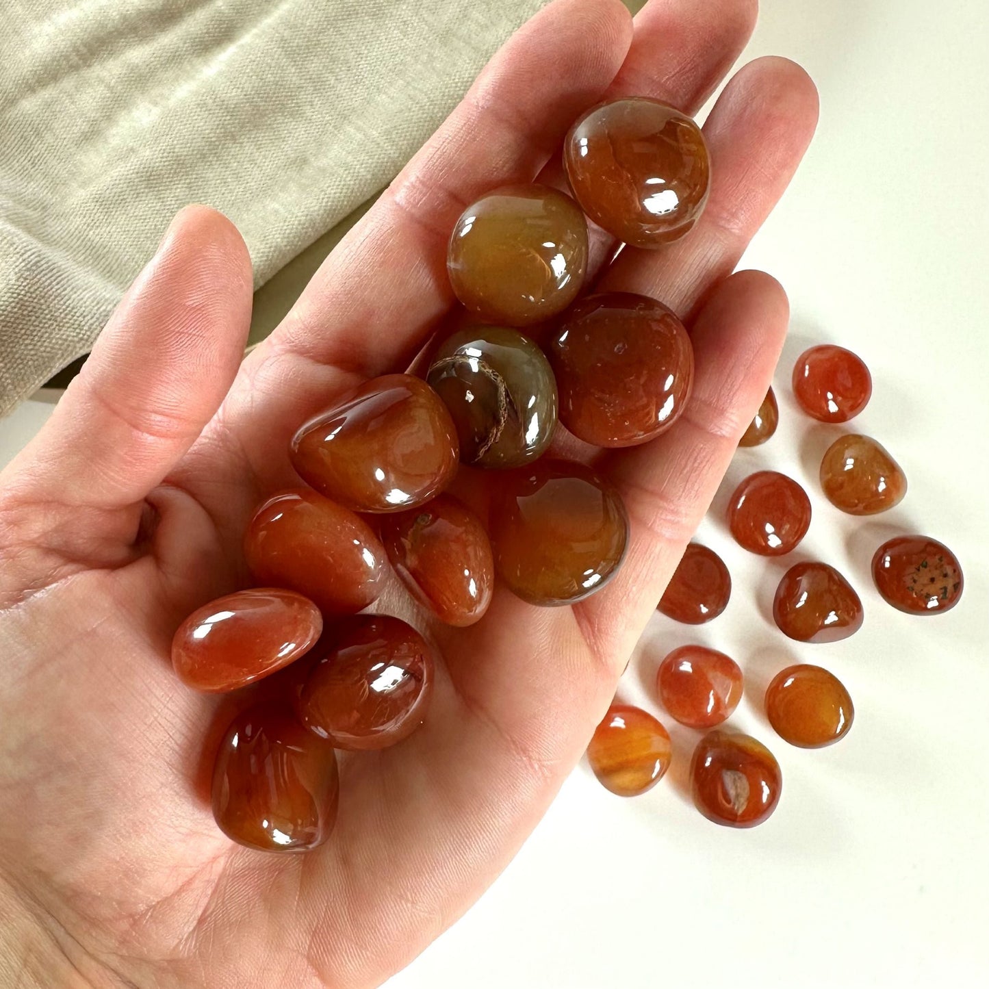 Hånd der holder glatte, polerede karneol lommesten med flere sten spredt på bordet i baggrunden.