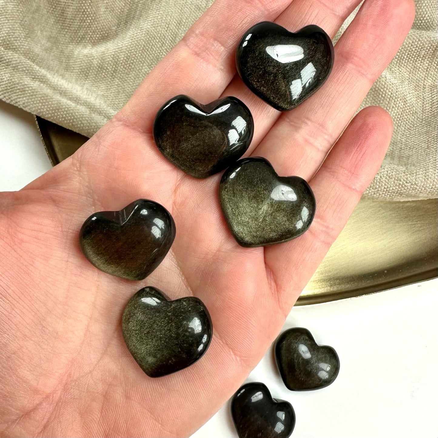 Hånden holder flere skinnende Guld Obsidian hjerter på en neutral baggrund.