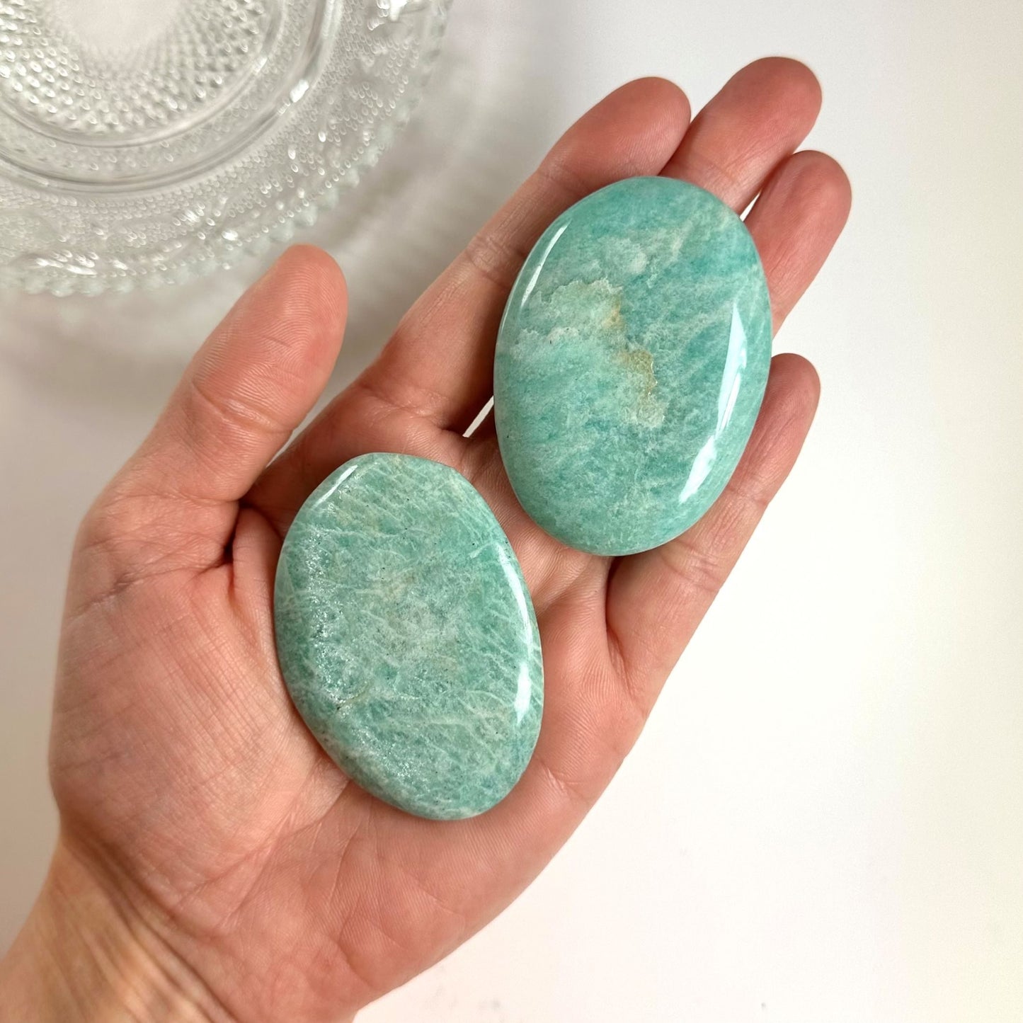 En hånd holder to glatte, ovale amazonit-sten på en lys baggrund, ved siden af en dekorativ glasskål.