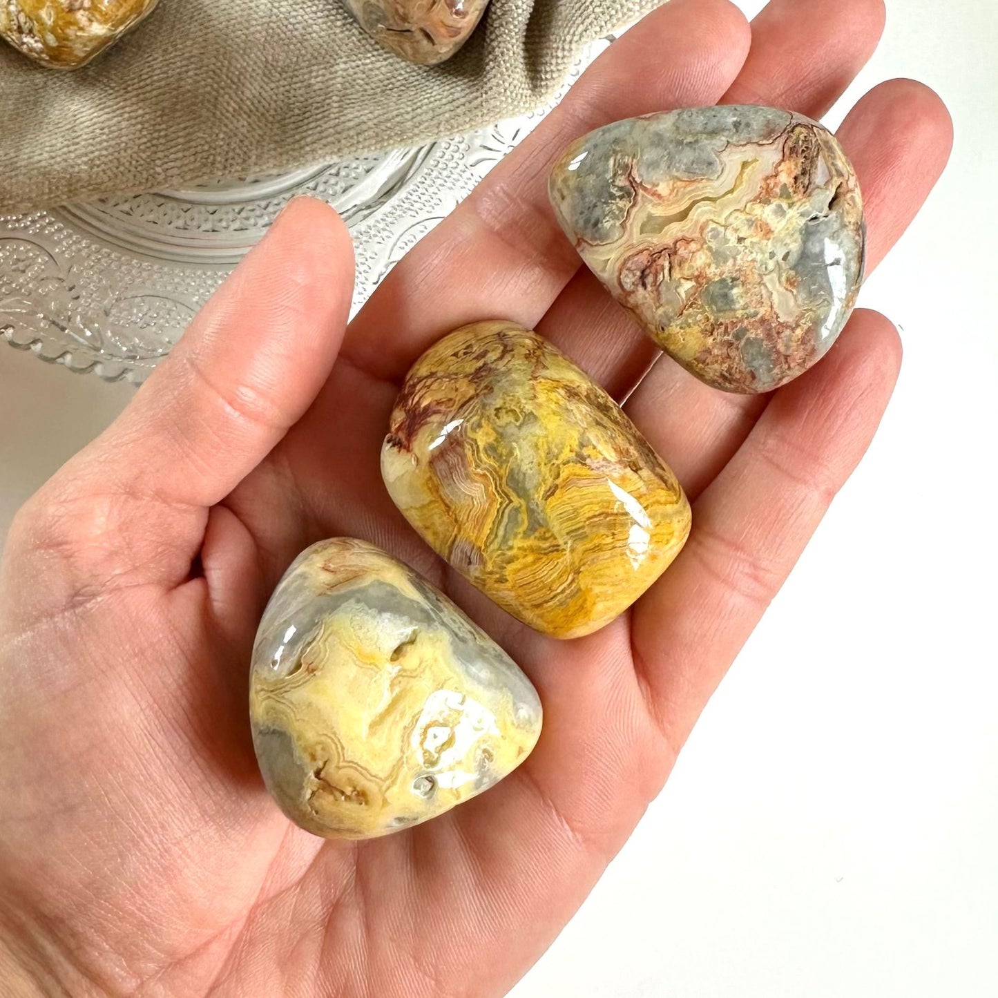 Hånd holder tre glatte Agat Crazy Lace XL sten med mønster i gule, grå og rødlige farver.
