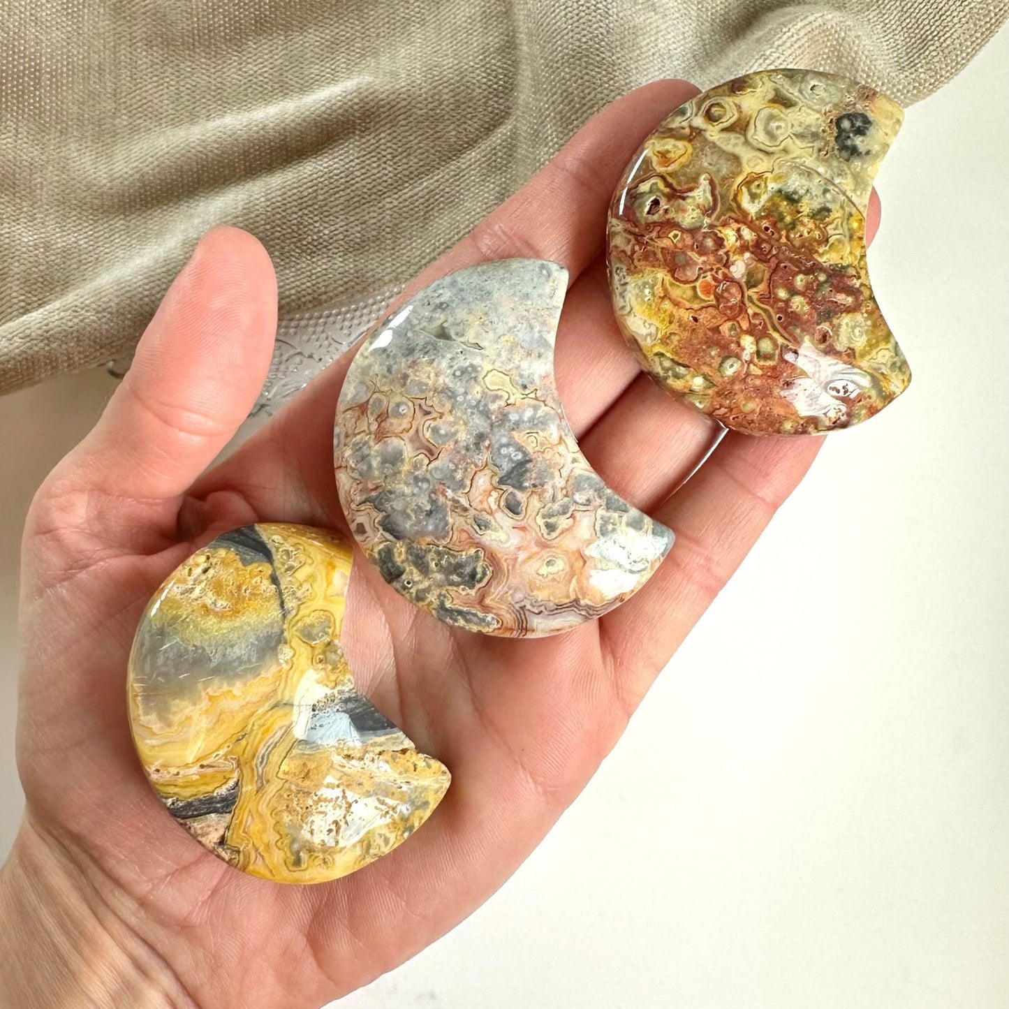 Tre agat crazy lace måner i forskellige farver ligger i en hånd, med beige stof i baggrunden.