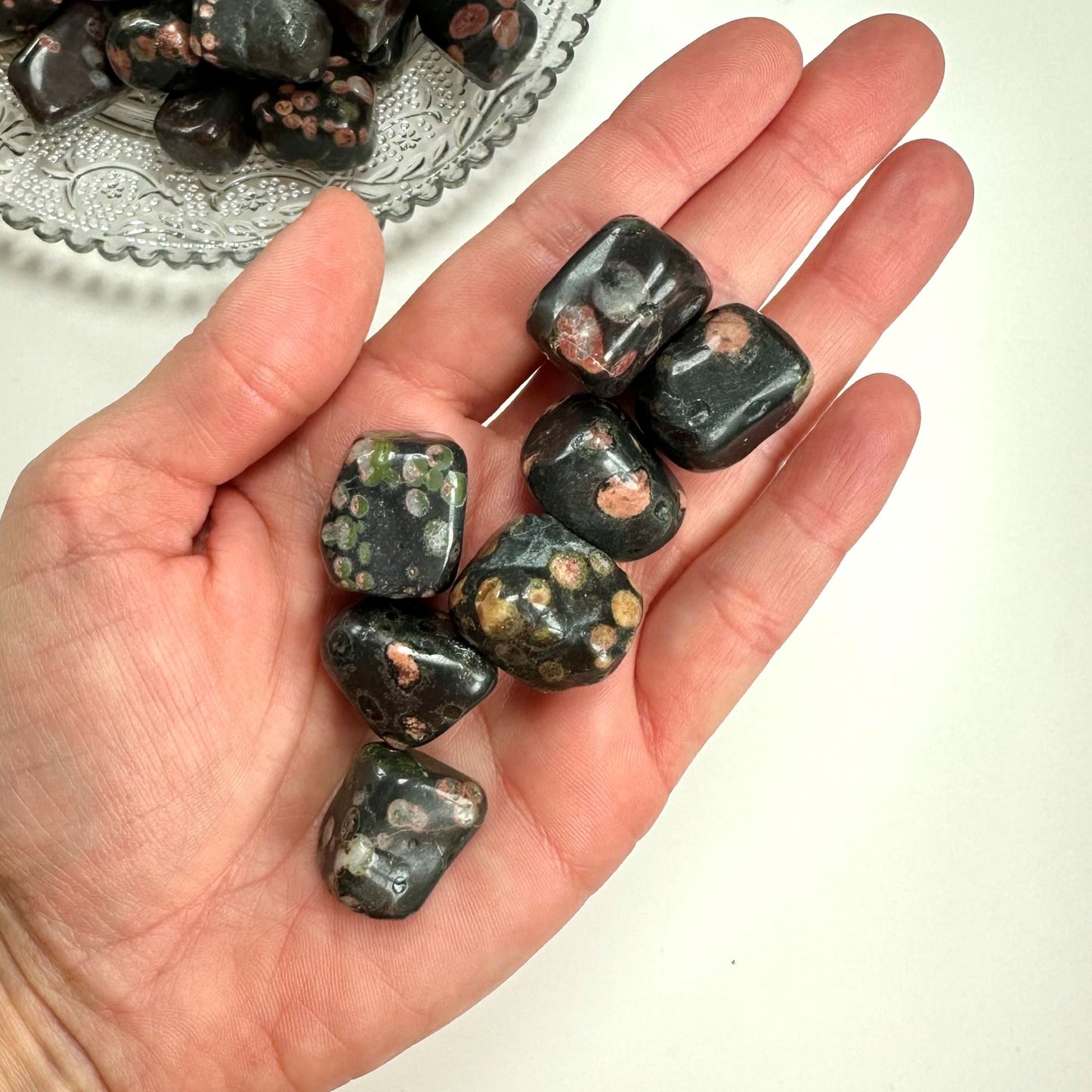 En hånd holder flere Jaspis flower lommesten med unikke mønstre og farver, en skål ses i baggrunden.