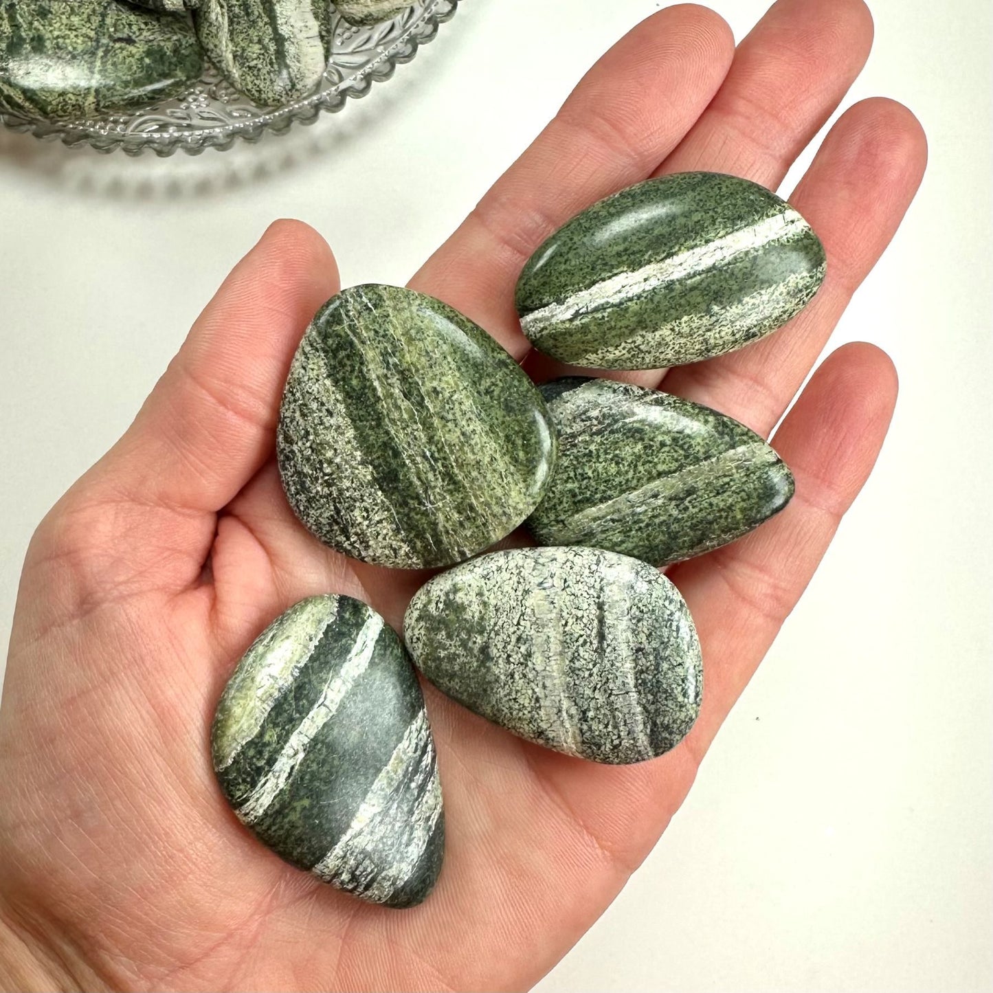 Hånd holder Serpentin Zebra lommesten med grønne og hvide linjer, flere på en tallerken i baggrunden.