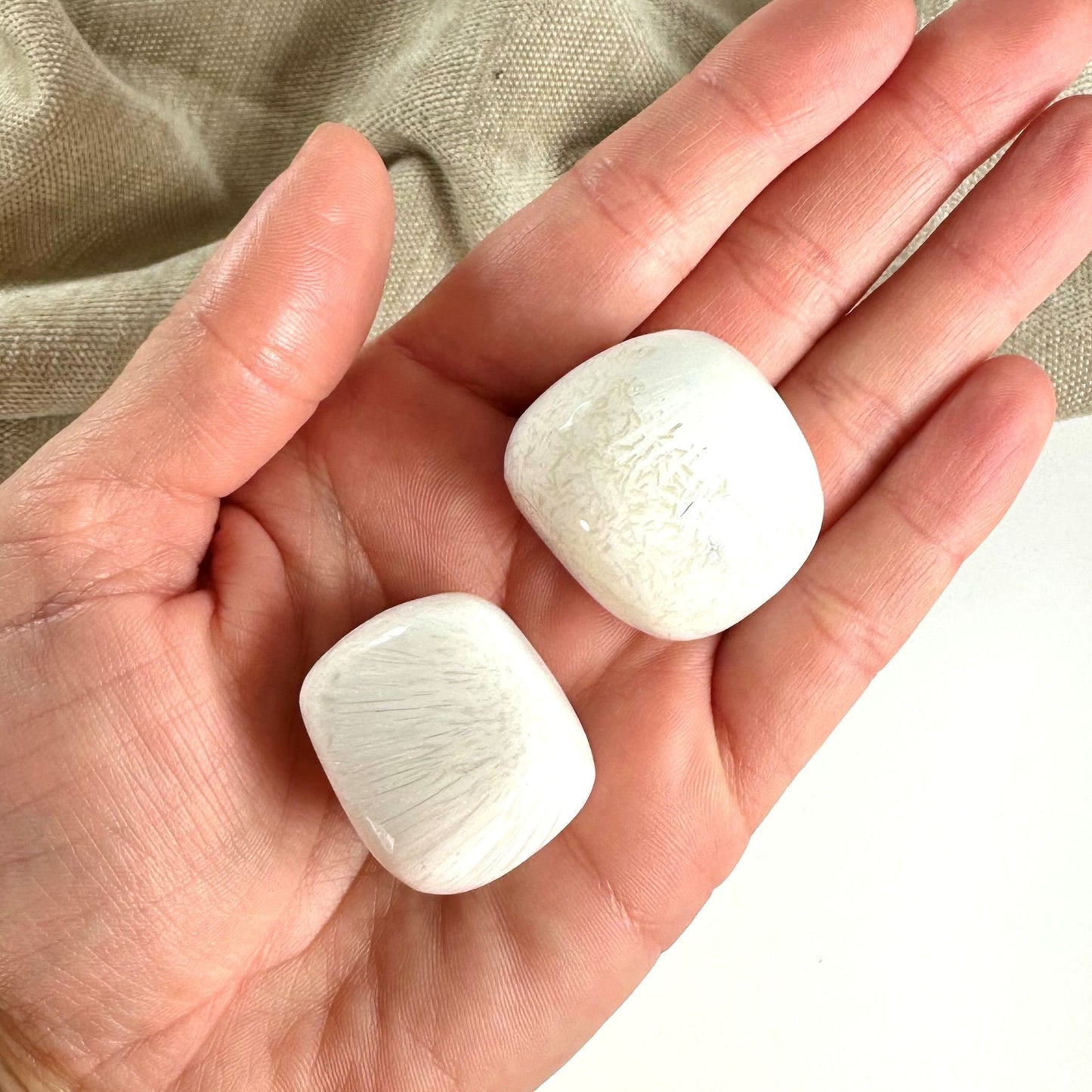 Hånd der holder to glatte, hvide scolesit jumbo sten på en lys baggrund.