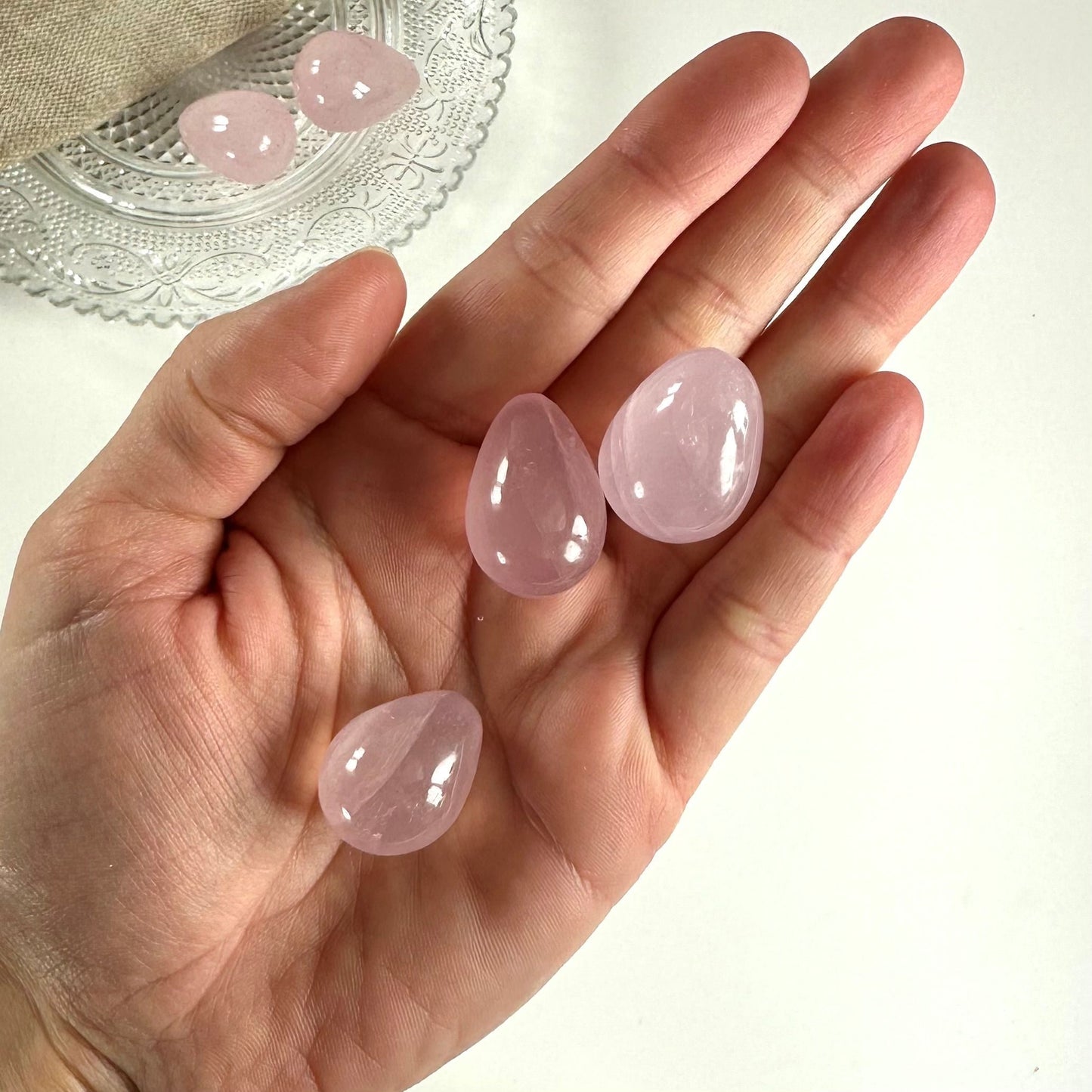 Hånd holder tre rosenkvarts æg, med to flere på en dekorativ tallerken i baggrunden.