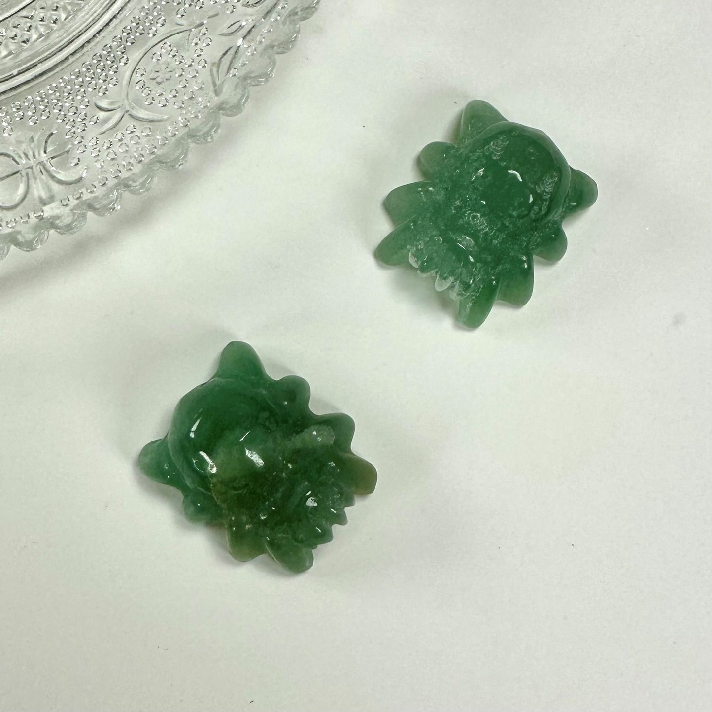 To grøn aventurin edderkopfiguriner på en hvid baggrund nær en dekorativ glasplade.