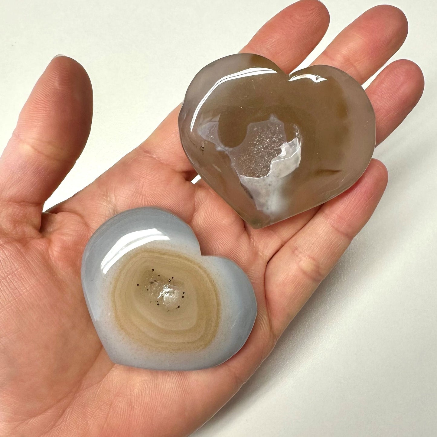 Hånd med to Agat natur hjerter, en brun og en grå, på en lys baggrund.