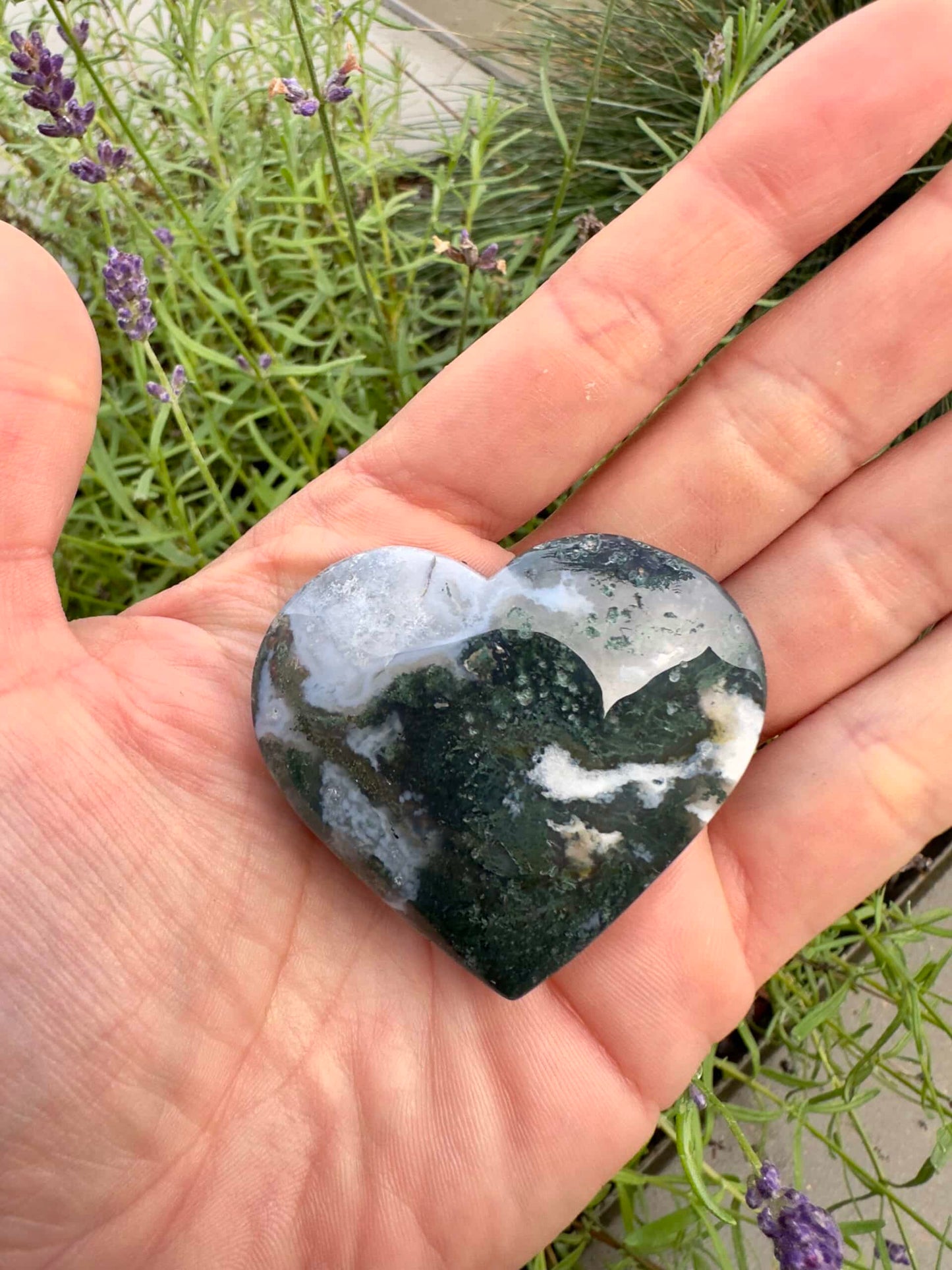 Hånd holder en Mos Agat hjerte sten foran grønne planter i baggrunden.
