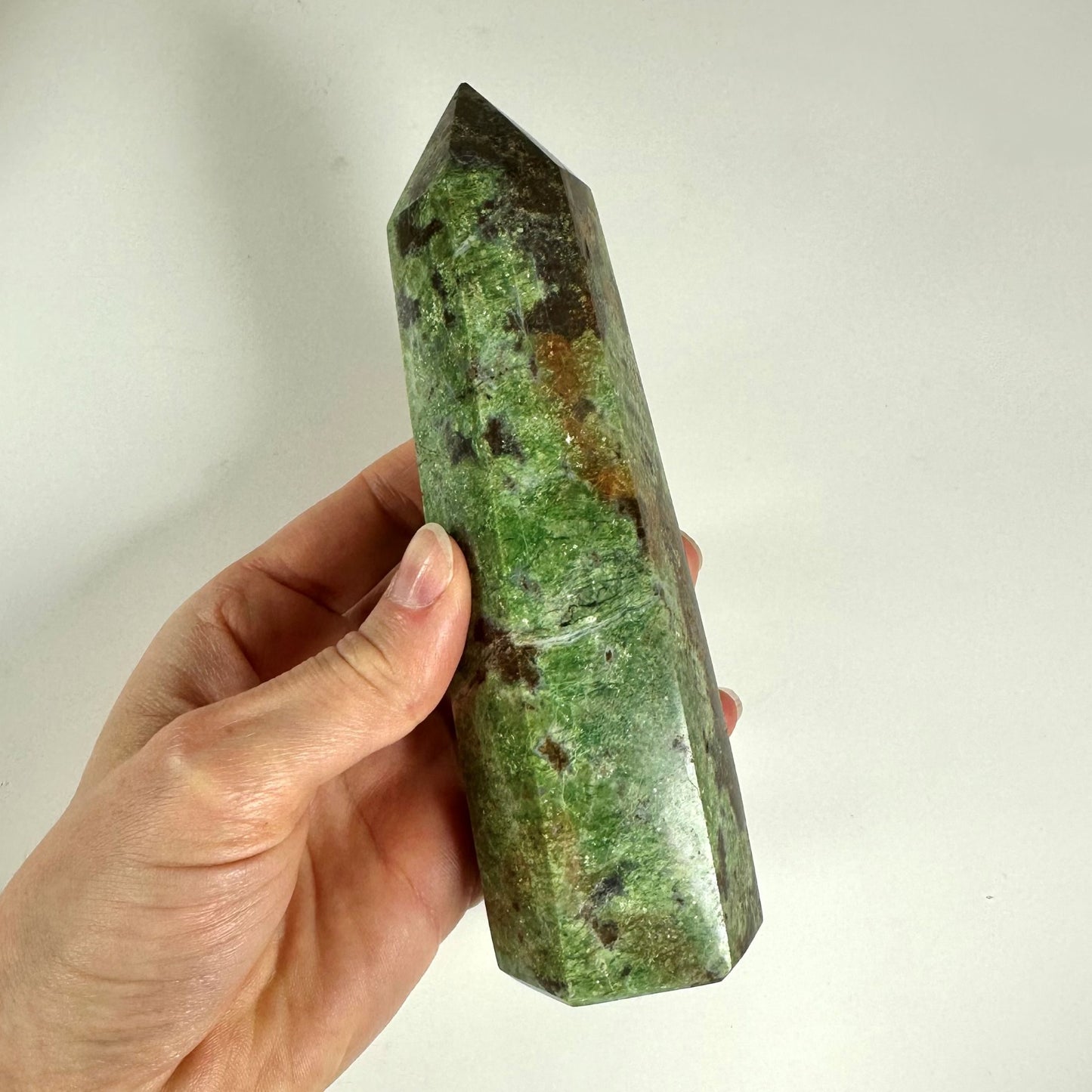 En hånd, der holder et poleret Chrysopras tårn med grønne og brune nuancer på en lys baggrund.