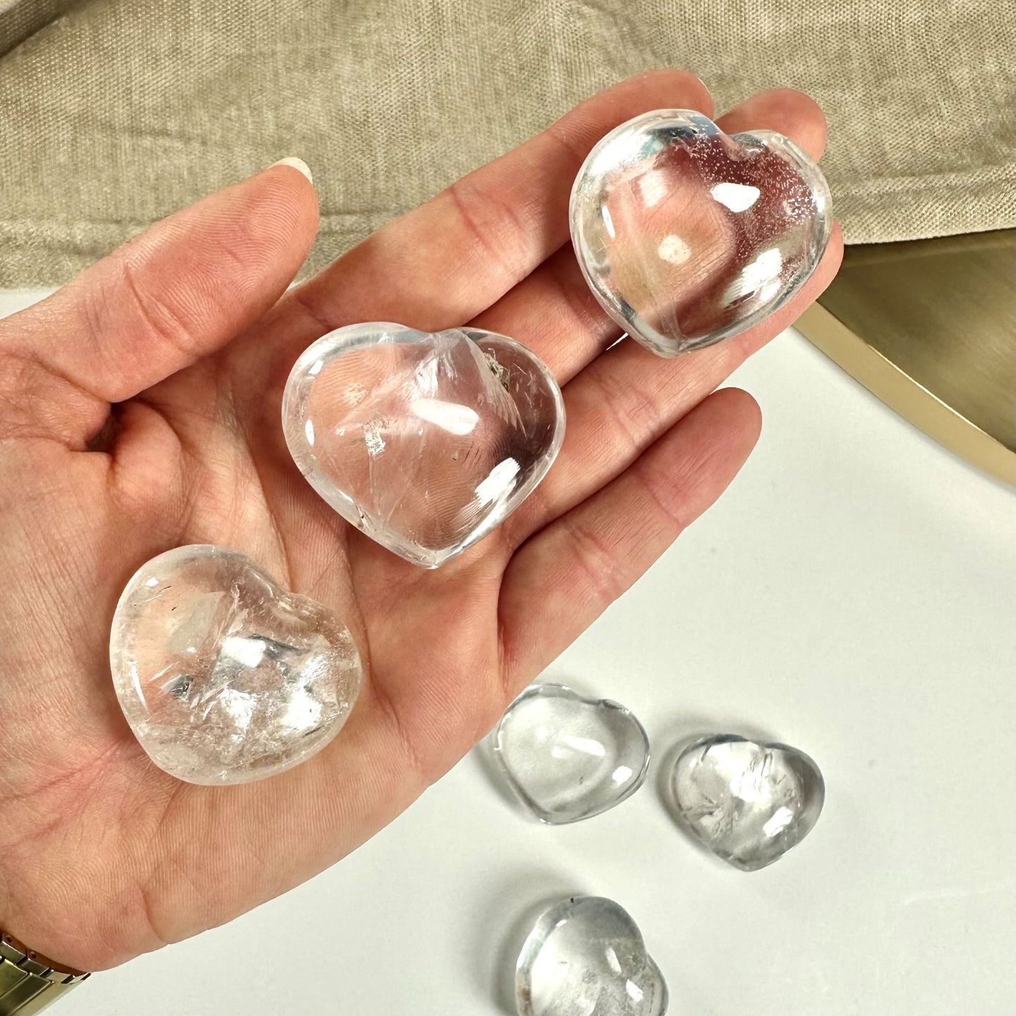 Hånd holder tre gennemsigtige bjergkrystal hjerter, med flere liggende på bordet.